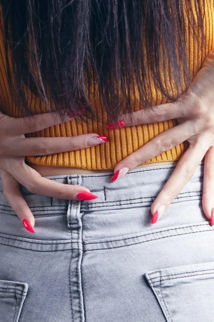 Person with long dark hair, wearing a mustard yellow ribbed top, has hands with long red painted nails placed on their lower back, above light gray jeans. Back Pain