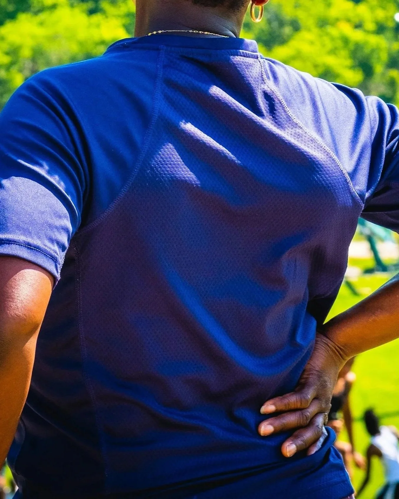 The back of a person wearing a blue athletic shirt with one hand on their hip, outdoors with green trees in the background. Sciatica