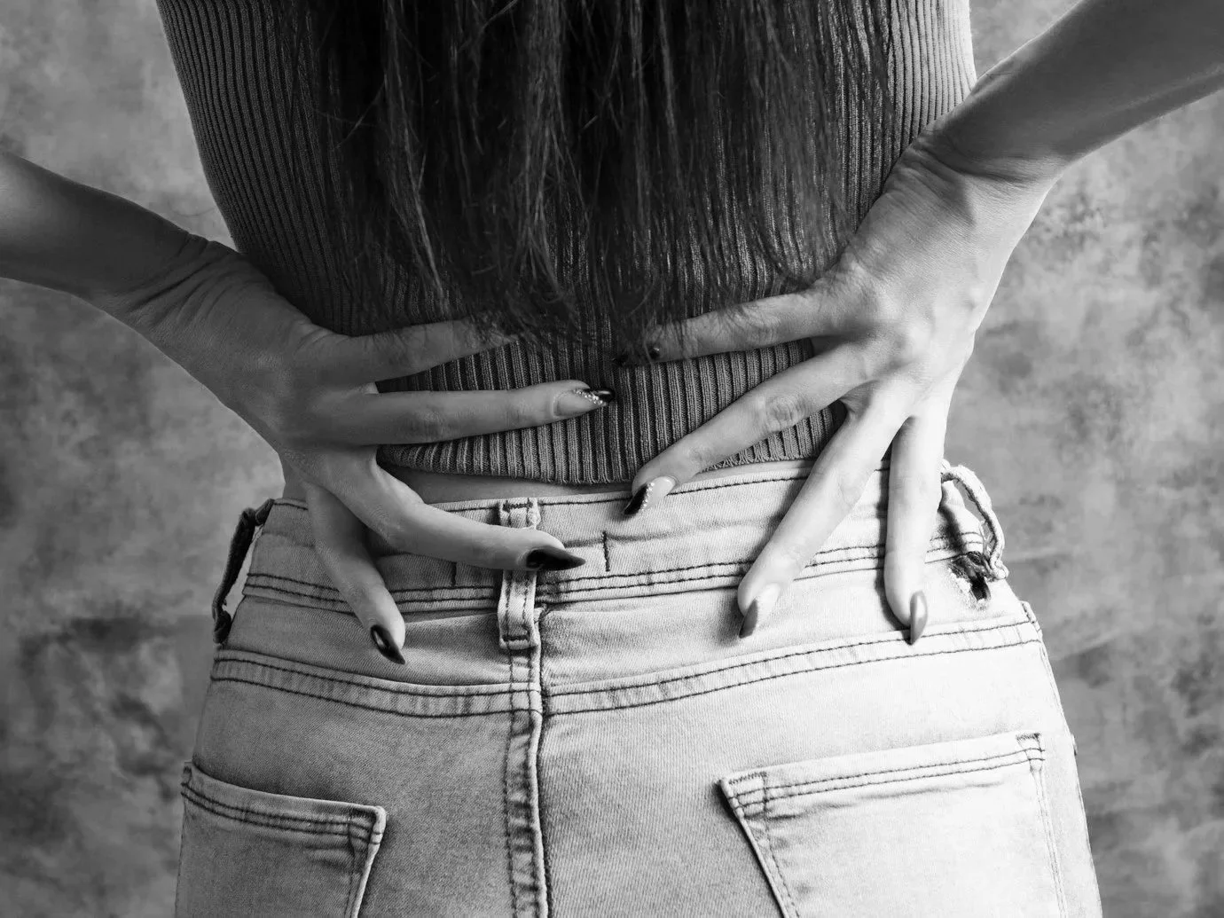 A black-and-white photo of a person standing with their back to the camera, wearing a ribbed top and light-colored jeans, with hands placed on their lower back and waist. back pain