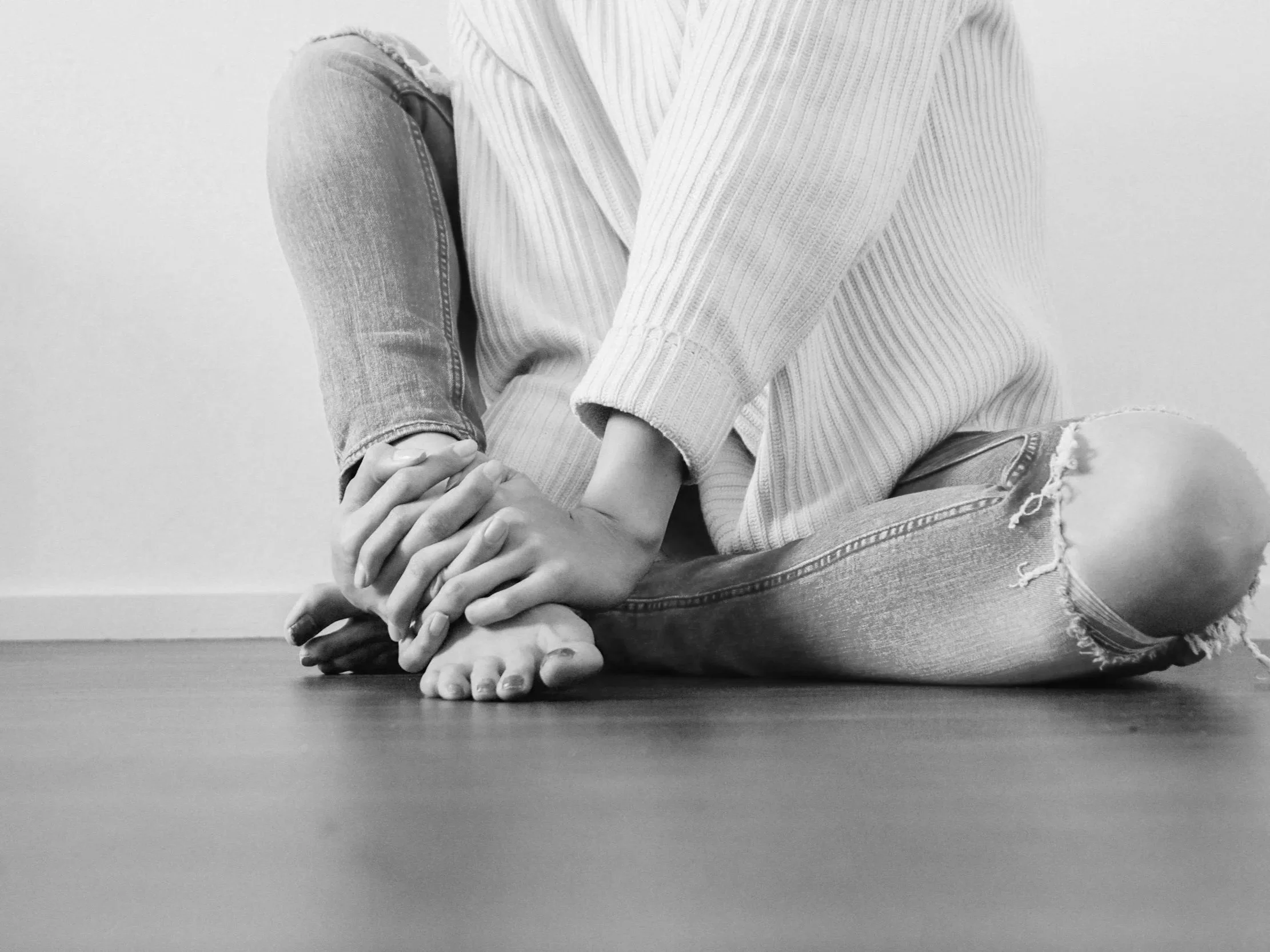 Black and white photo of a person seated cross-legged with hands clasped on their knees, wearing ripped jeans and a striped sweater Foot pain Orthotics