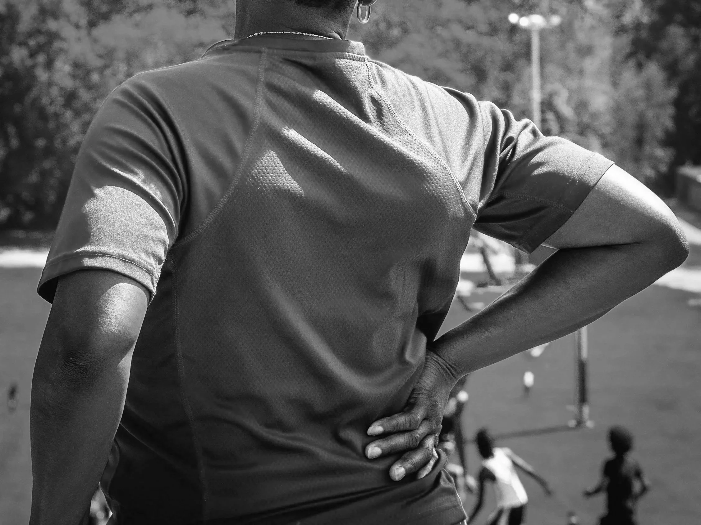 Close-up of a person wearing a short-sleeve athletic shirt, with one hand on their hip, standing outdoors on a sunny day with blurred trees and a lamppost in the background. Sciatica