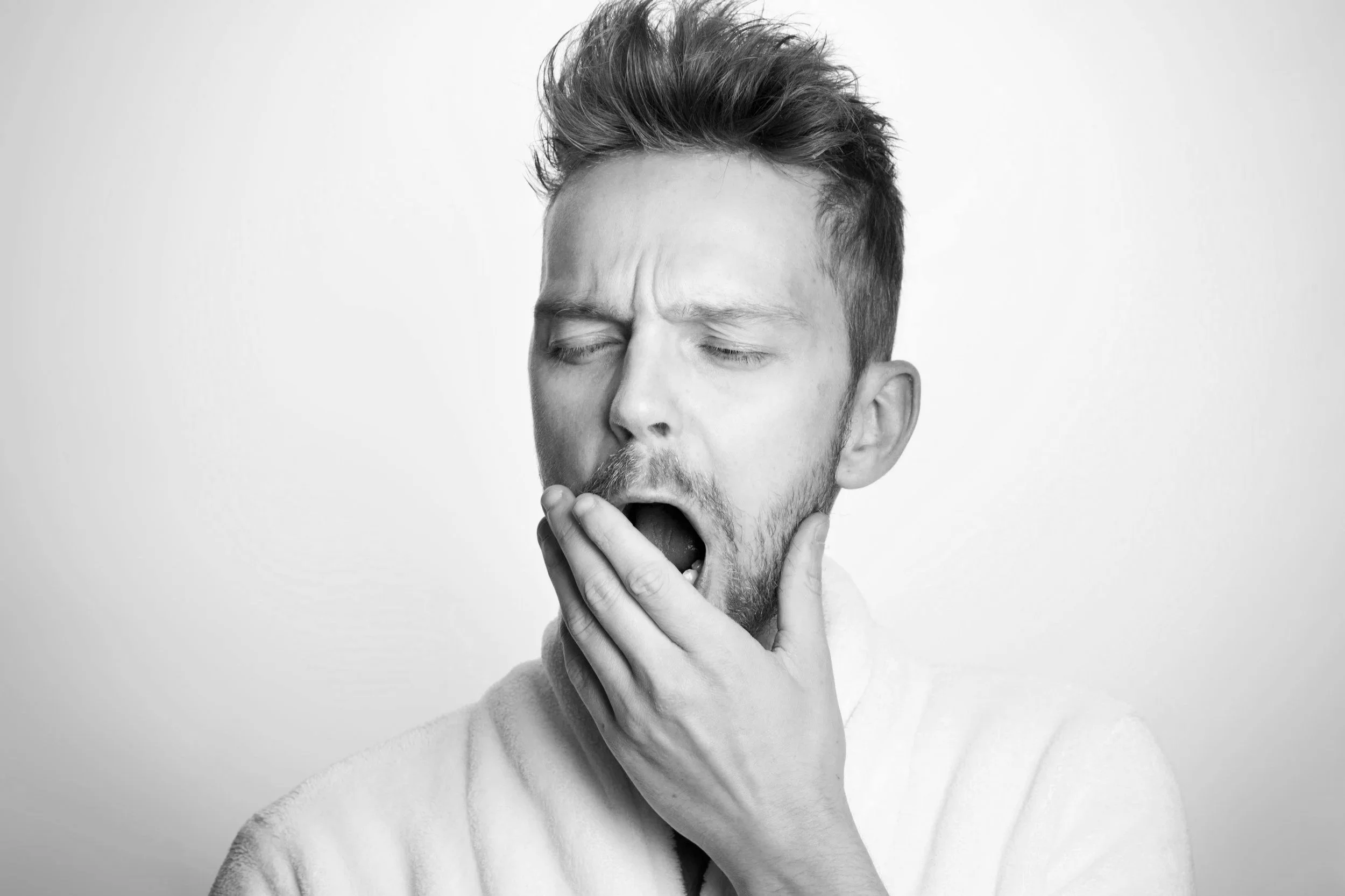 A black and white photo of a young man with styled hair, closed eyes, and his mouth open, appearing to yawn or cough, with one hand on his chin. TMJ pain