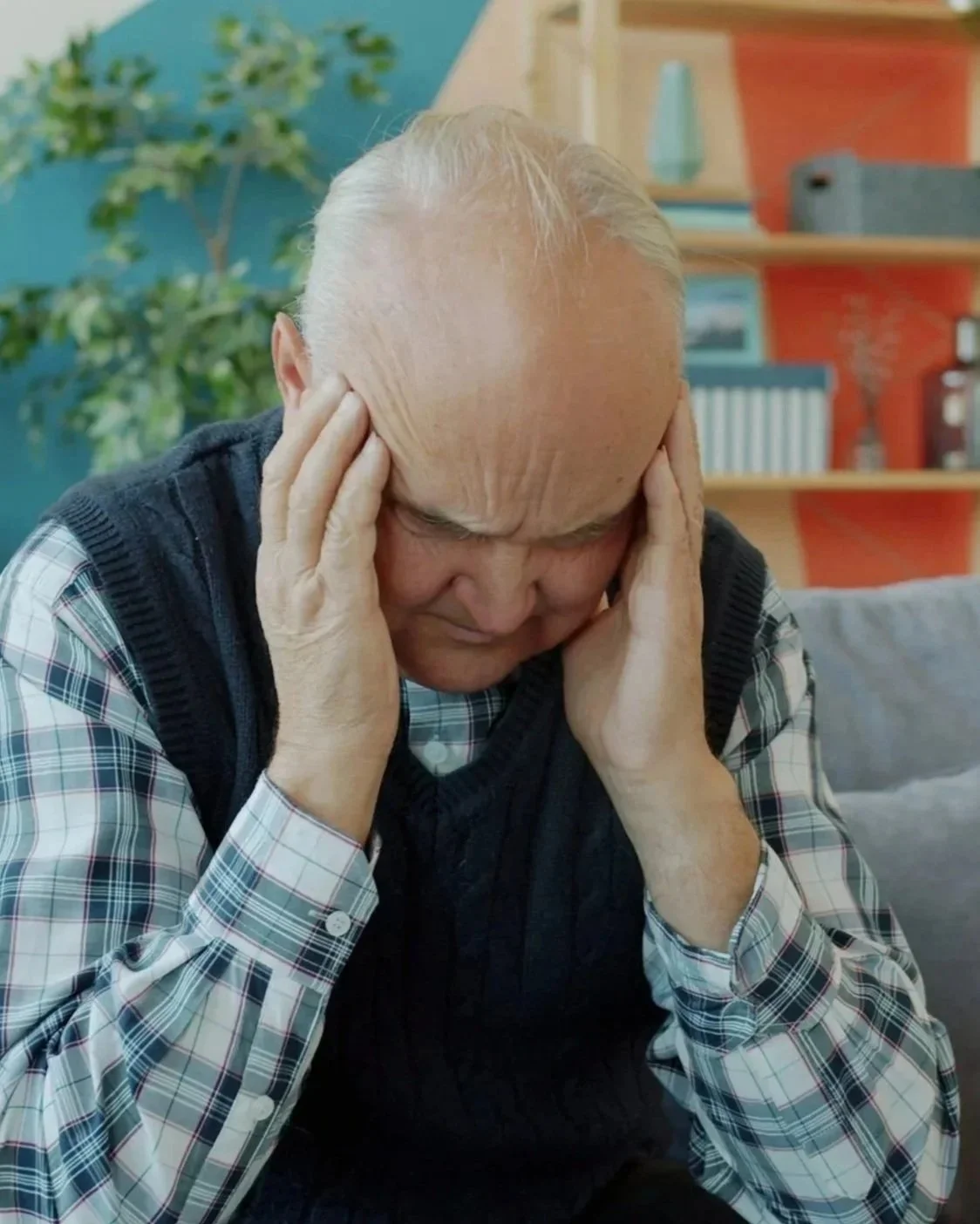 An elderly man with gray hair, wearing a plaid shirt and a navy blue vest, is holding his head with both hands and appears to be experiencing pain or discomfort indoors, with a colorful background including a bookshelf, a plant, and some decorative objects.