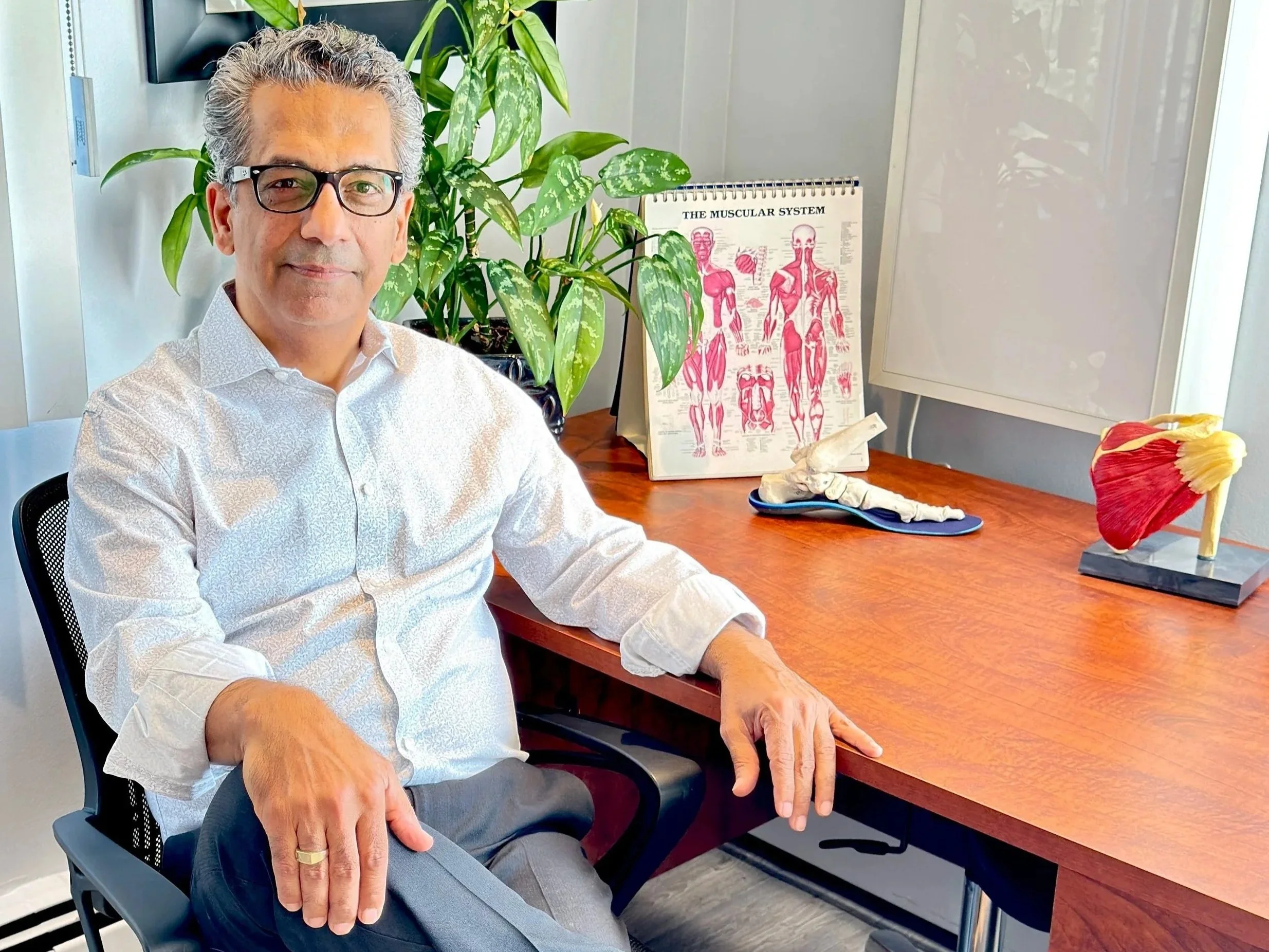 A man with gray hair, glasses, and a light-colored patterned shirt sitting at a wooden desk. Behind him is a green plant, a chart of the muscular system, and a whiteboard. Dr. Babak Missaghi