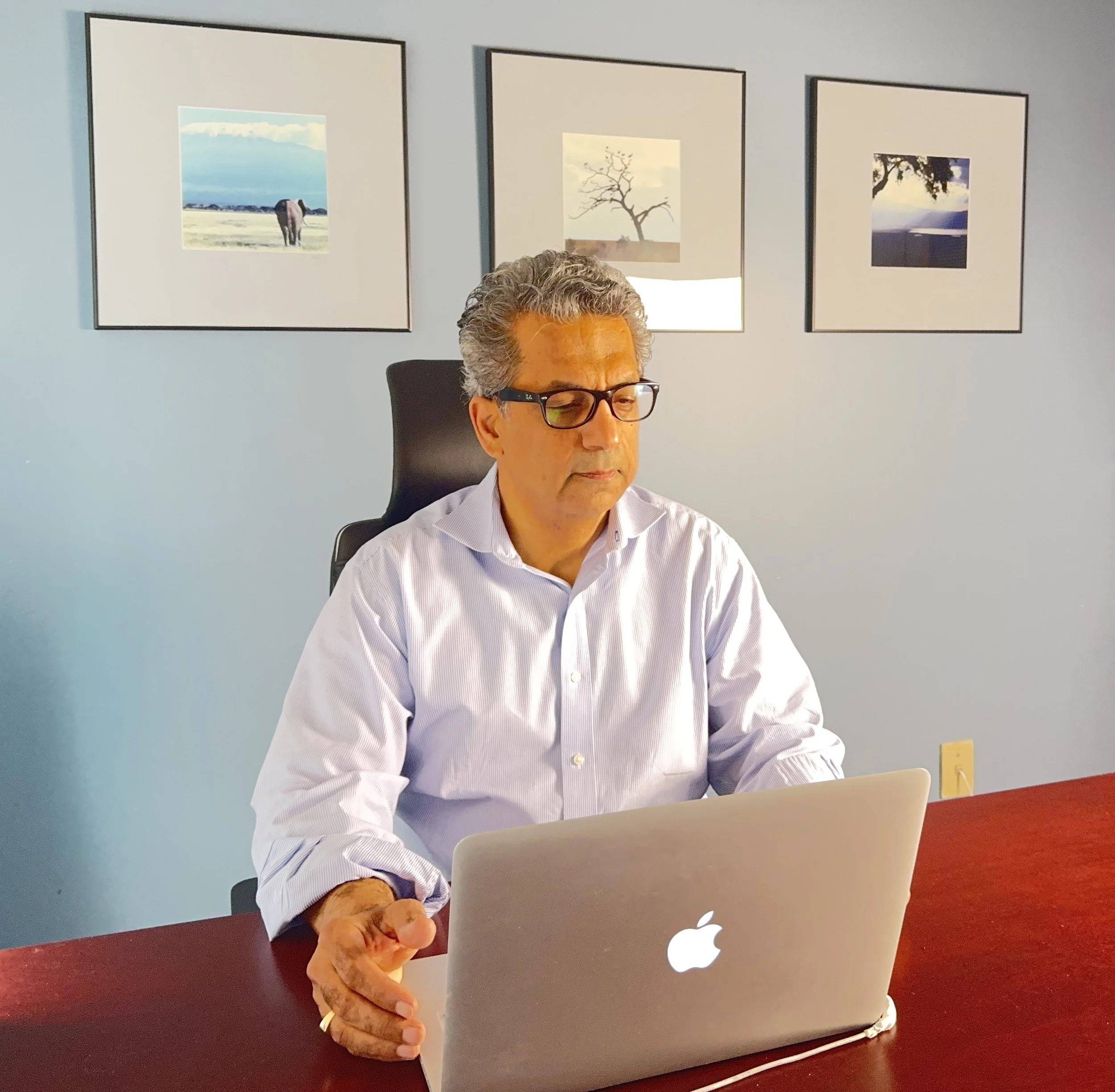 An older man with gray hair and glasses working on a laptop at a desk in an office, with three framed landscape photographs on the light blue wall behind him. Dr. Missaghi during virtual consultation