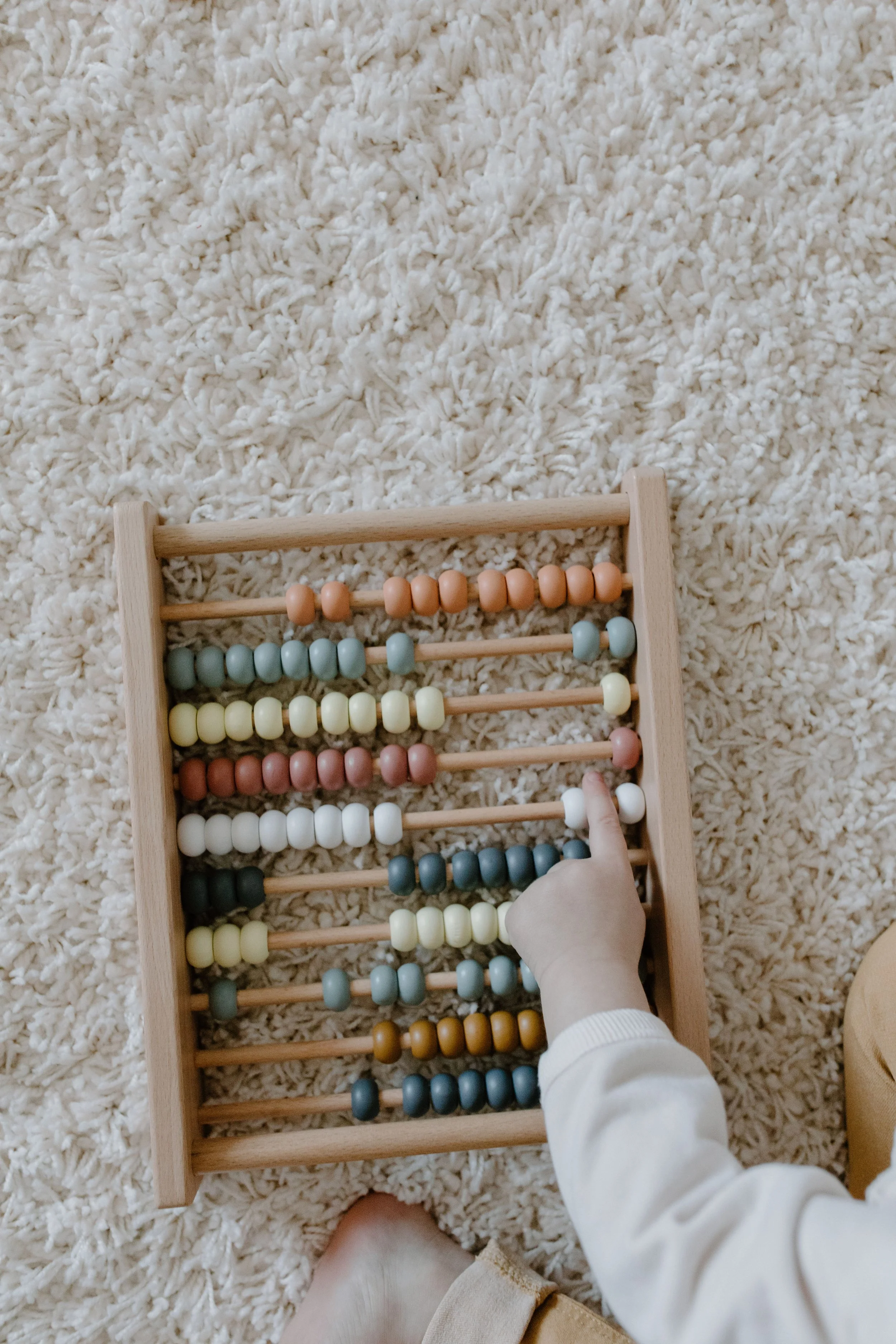 Une main d'enfant utilisant un boulier en bois avec des perles de différentes couleurs, posé sur un tapis blanc épais.