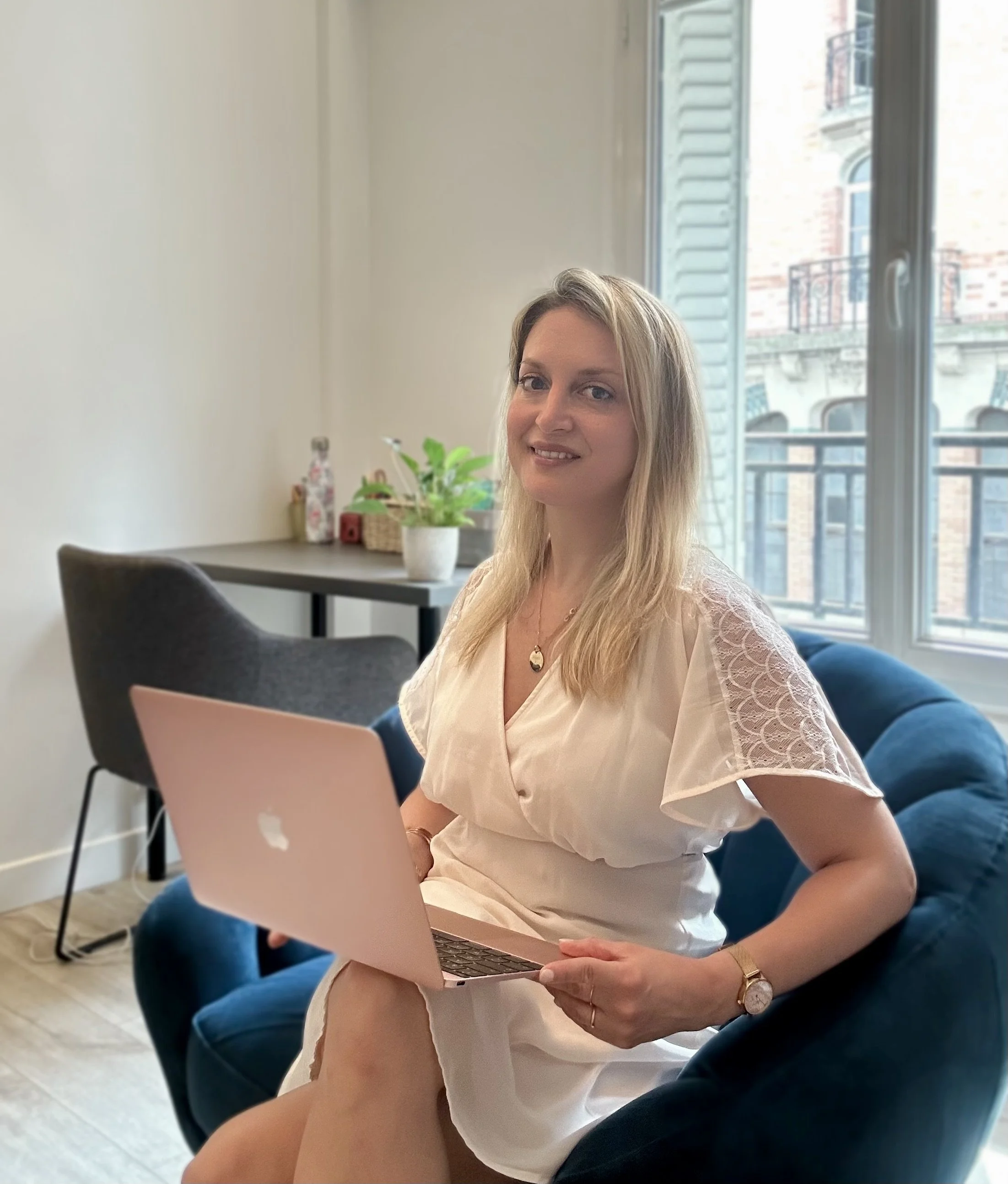 Une femme blonde assise sur un canapé en train de travailler sur un ordinateur portable rose, dans une pièce lumineuse avec une grande fenêtre et un balcon visible derrière elle.