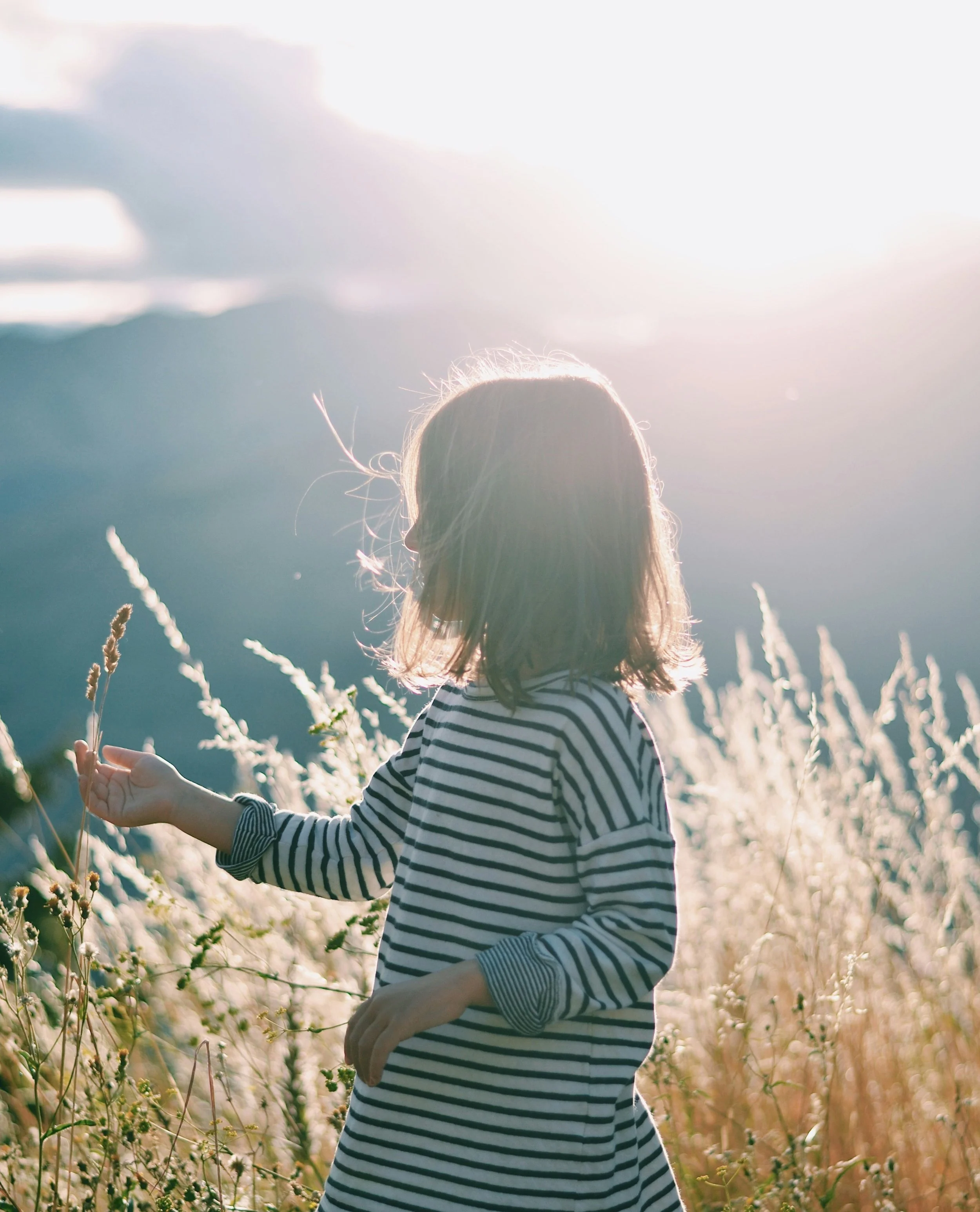 Une jeune fille avec des cheveux longs portant un pull rayé noir et blanc, dans un champ de plantes avec des fleurs pâles, ensoleillé avec un ciel lumineux et quelques nuages.