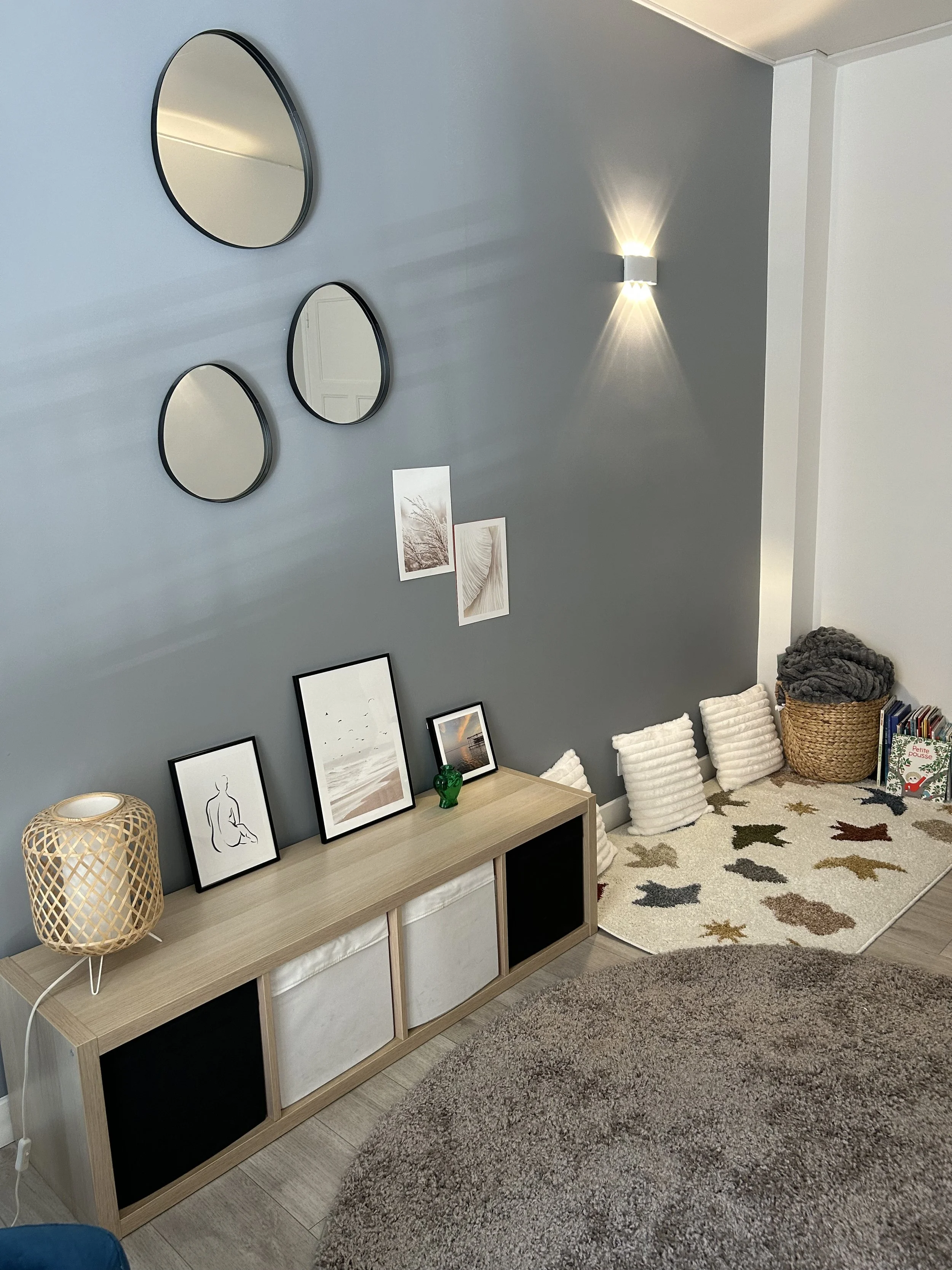 Une pièce de salon ou de chambre avec un meuble en bois clair contenant des boîtes de rangement blanches et noires, des cadres avec des illustrations minimalistes, un panier en osier avec des couvertures douces, un tapis moelleux avec des motifs étoilés, un mur gris avec des miroirs ronds et des œuvres d'art, et un espace avec des coussins rembourrés blanc et une petite bibliothèque avec des livres.