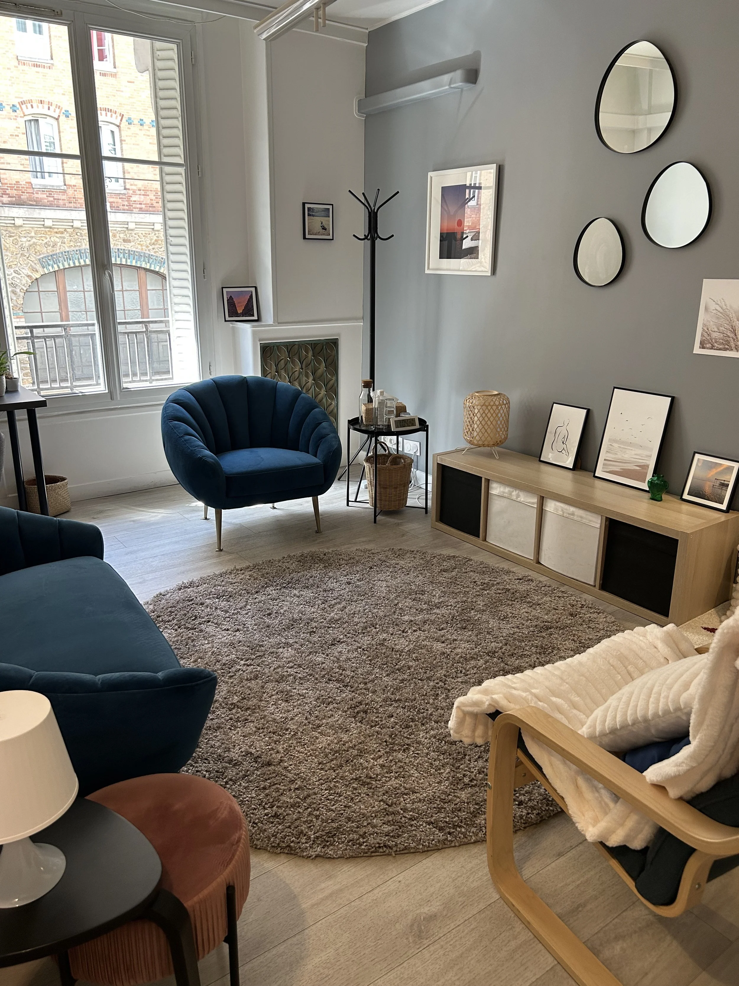 Salon intérieur avec un fauteuil bleu foncé, un tapis rond beige, une étagère en bois avec des cadres et une lampe en osier, un portant à vêtements, un canapé et un coin avec plusieurs miroirs ovales au mur.