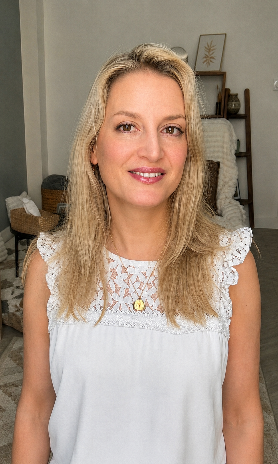Portrait de la psychologue Nathalie Soulié-Pain ; jeune femme blonde souriante portant une robe blanche avec des détails en dentelle, dans un intérieur décoré avec des plantes et des œuvres d'art.