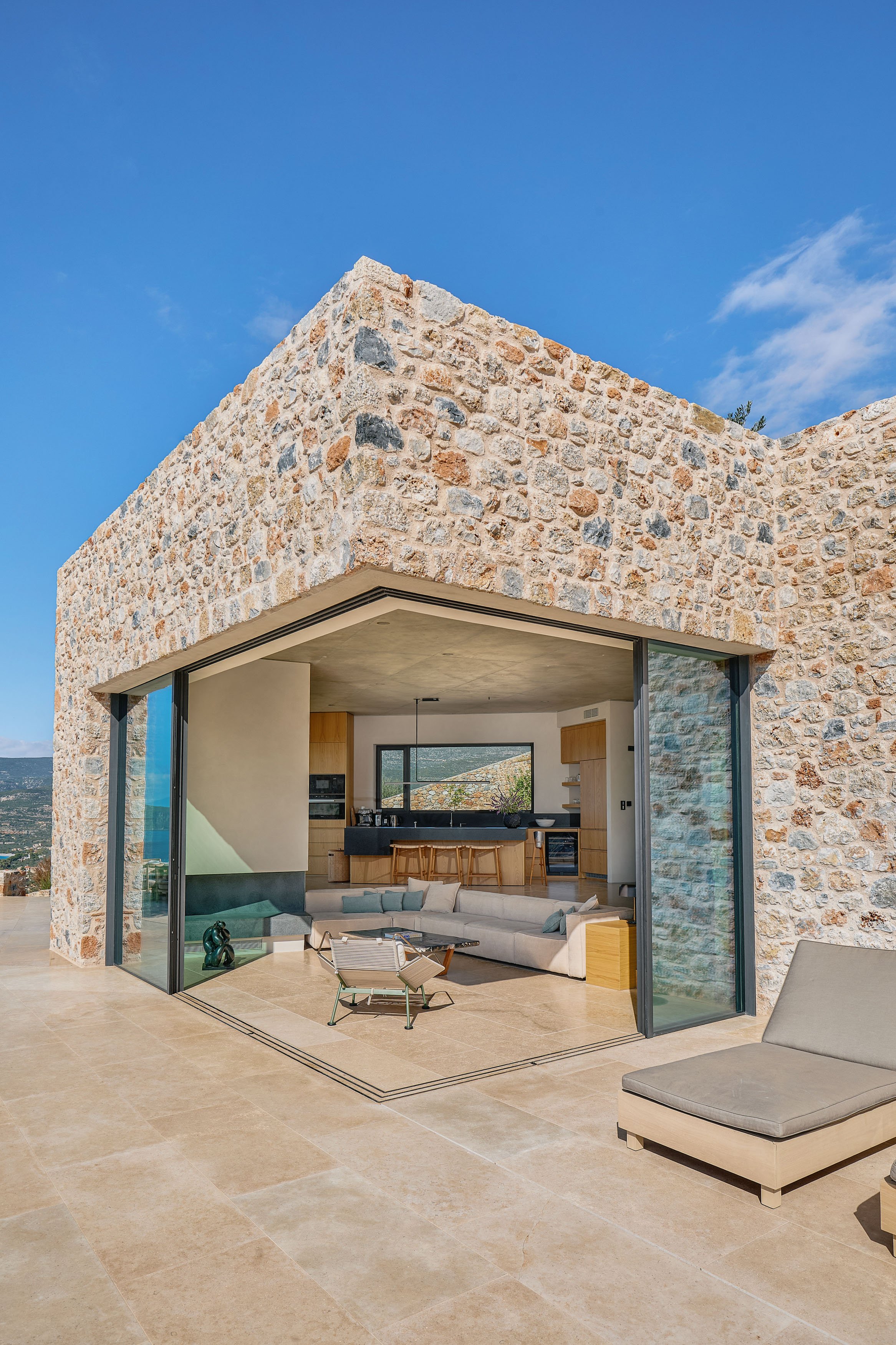 Modern house with stone exterior, open living area with glass walls, beige tiled floor, cream sectional sofa with pillows, small tray table, and lounge chair outdoors, clear blue sky, and mountain view in the background.