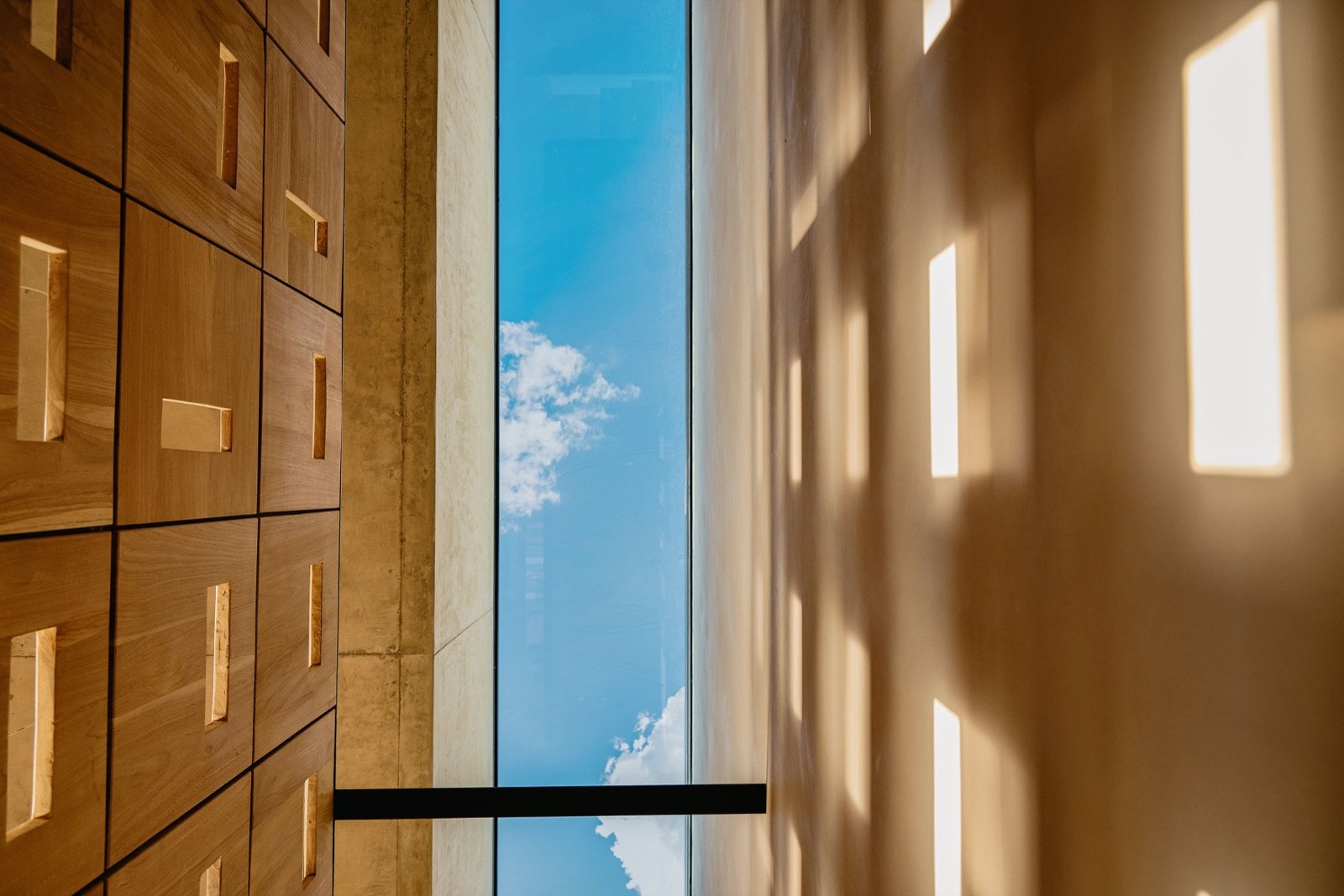 Interior view of a modern space with wooden cabinetry on the left, a wide open area with natural light streaming through rectangular openings in the ceiling, and a view of the sky with clouds visible through a large window or opening at the far end.