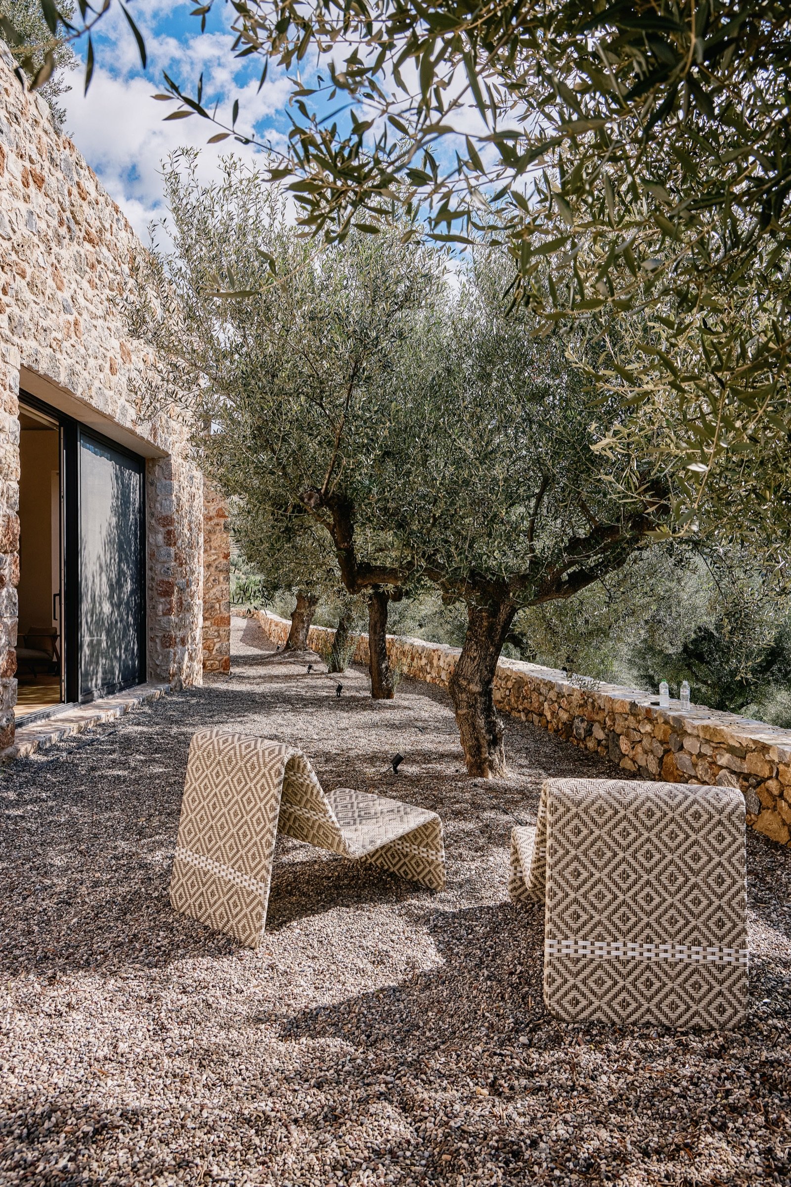An outdoor patio area with gravel ground and modern patterned chairs. Several trees with thick trunks and olive green leaves are along a stone wall, with a stone house and window to the left. The sky is partly cloudy.