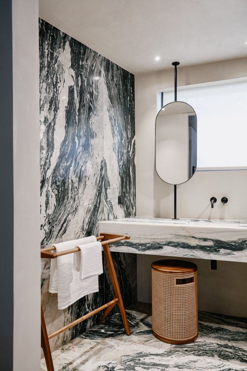 Modern bathroom with a black and white marble wall and floor, a white marble sink, a tall oval mirror, a wooden towel rack with white towels, and a small woven laundry basket.