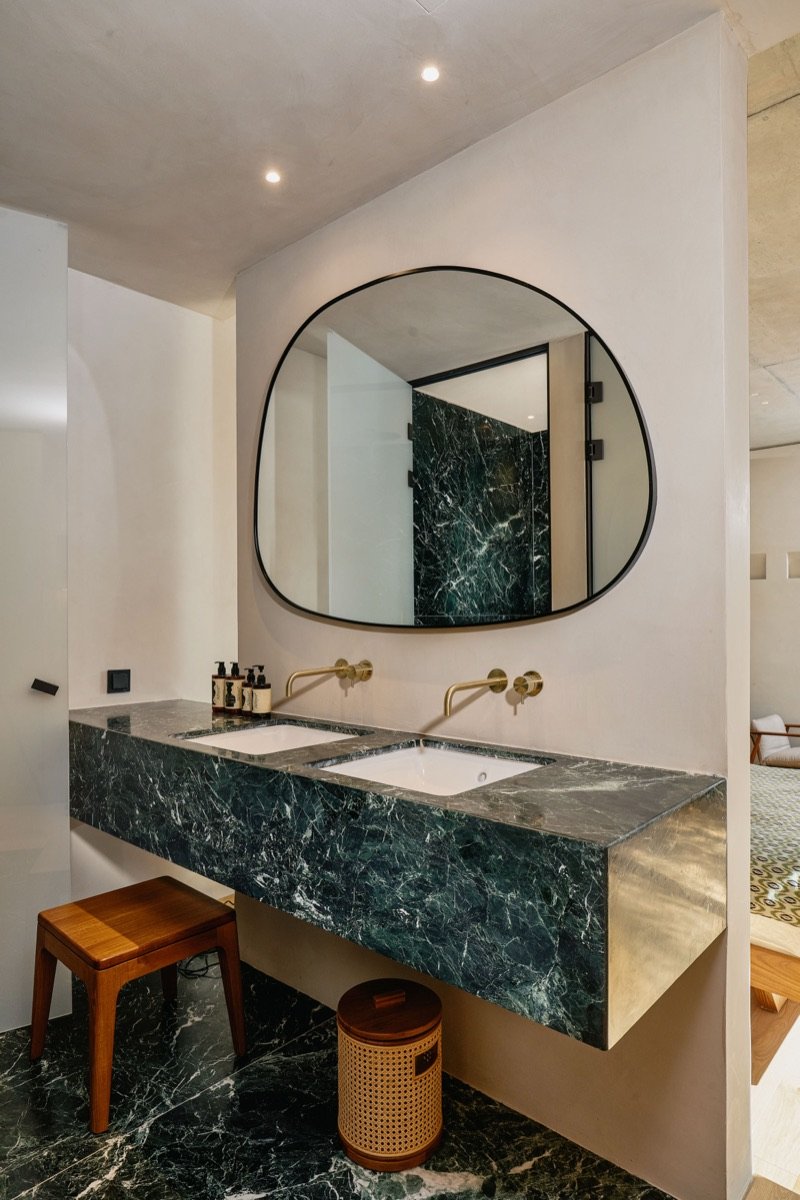 Modern bathroom with double green marble sinks, gold faucets, a large irregular-shaped mirror, and minimal decor.