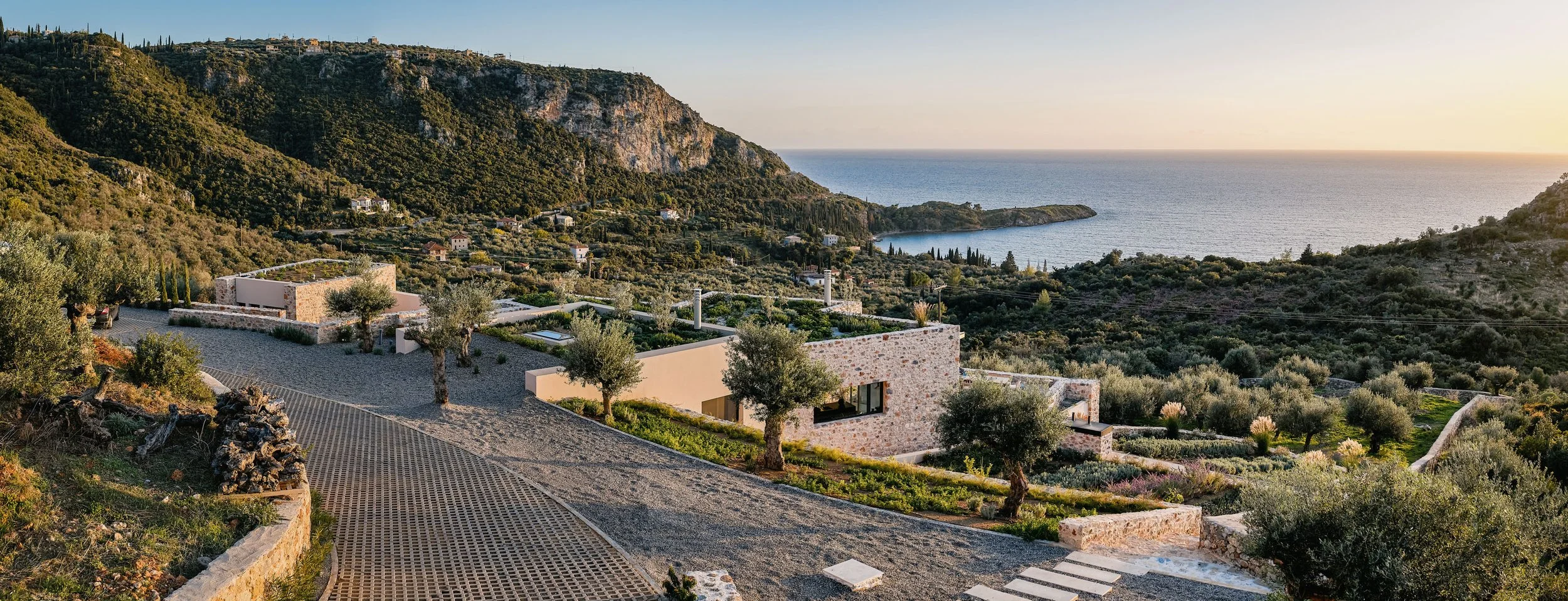 Modern house with stone exterior walls and large windows surrounded by olive trees on hillside overlooking the sea with hills in the background