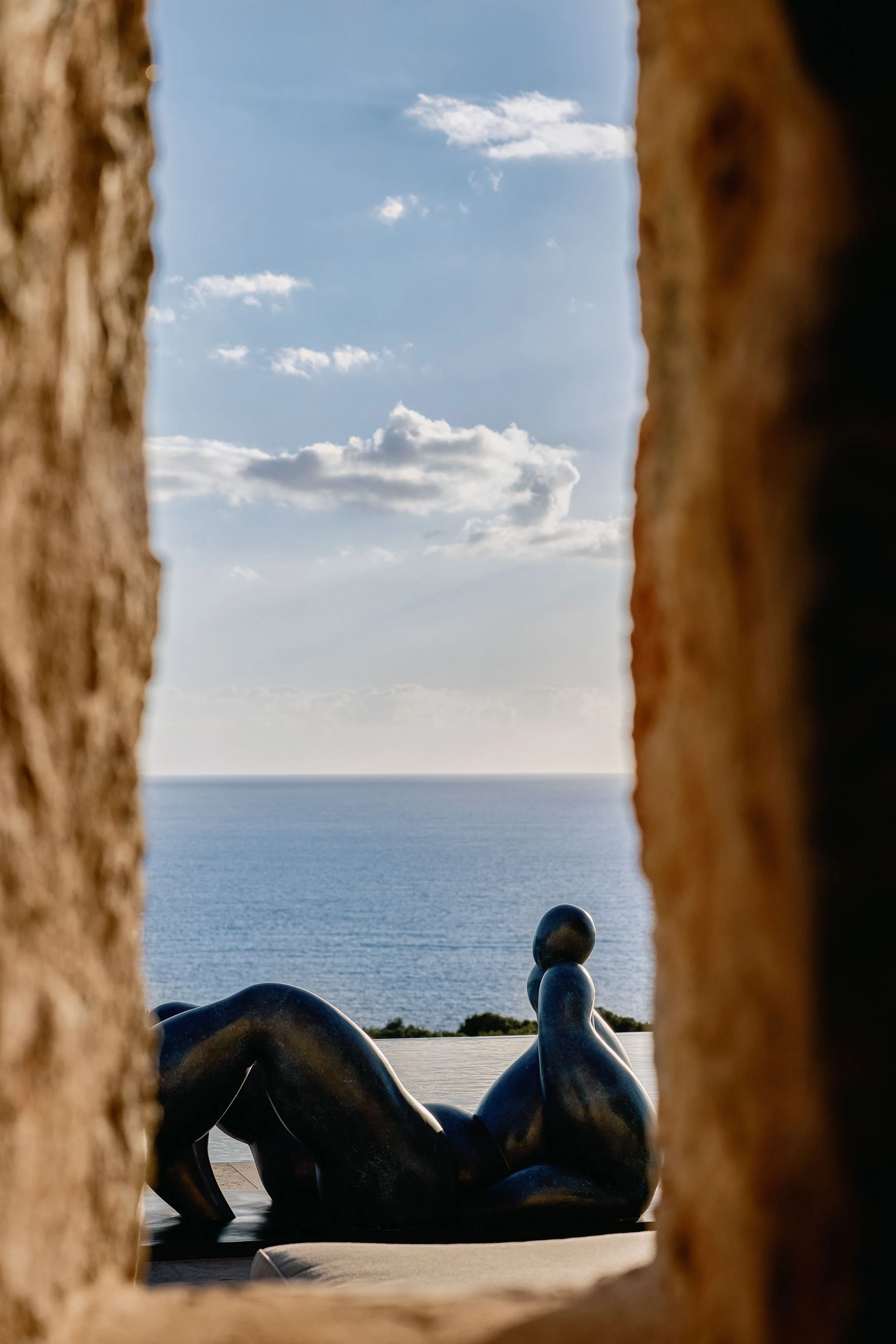 View of the ocean with a partly cloudy sky, seen through a narrow gap between two stone or brick structures. In the foreground, there are abstract black sculptures.