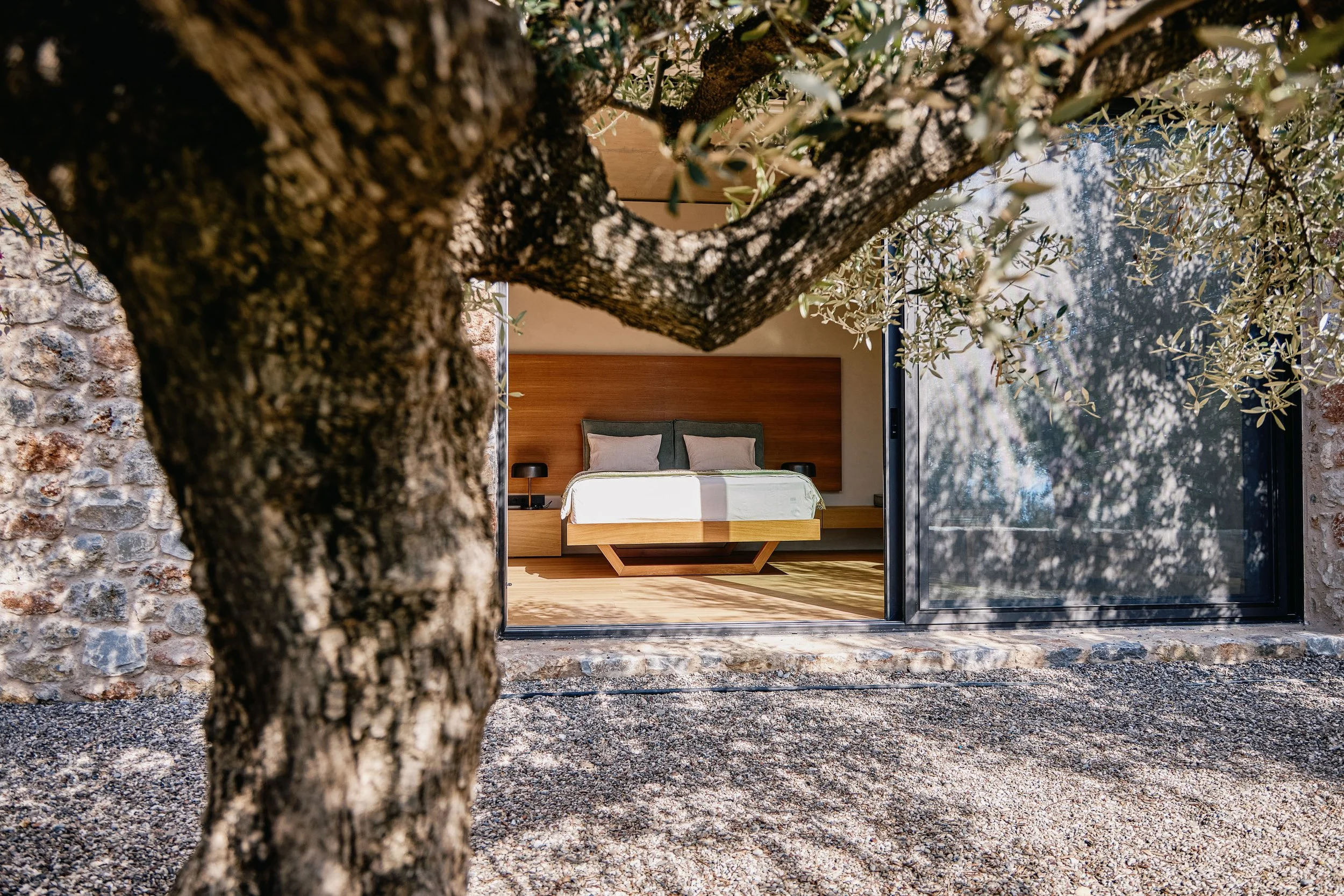 A modern bedroom visible through a large glass window, with a bed, pillows, and wooden headboard, surrounded by a stone wall and trees outside.