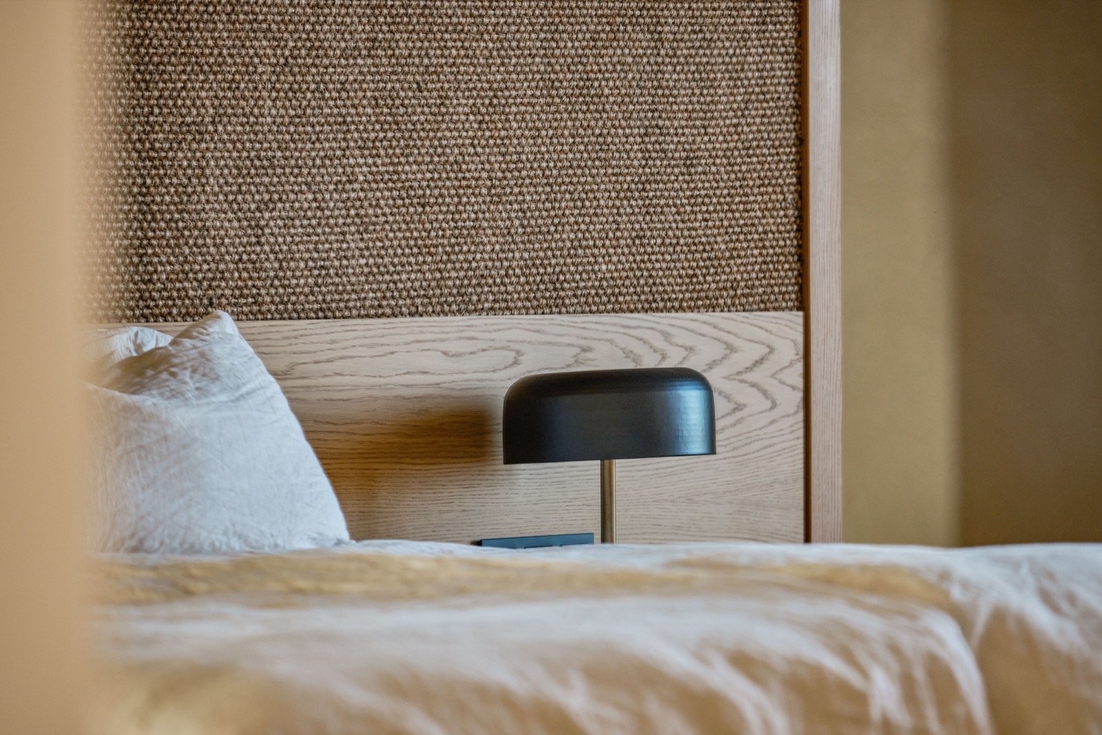 A bedroom with a bed, white bedding, a headboard with wood and fabric, a black bedside lamp, and beige walls.