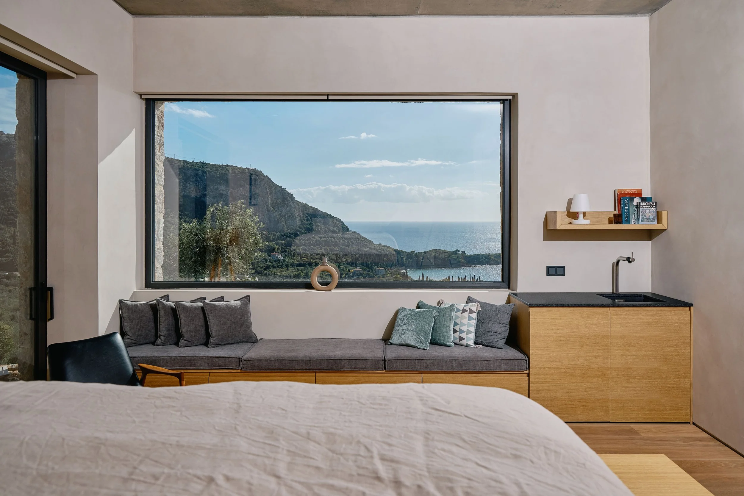 A modern living room with a large window offering a scenic view of hills and water. The room includes a couch with cushions, a wooden side cabinet with books and a lamp, and a portion of a bed is visible in the foreground.