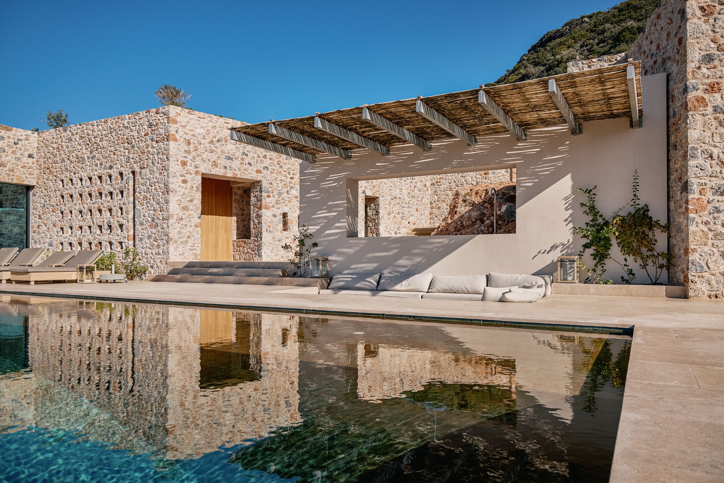 Modern stone and stucco house with a pool, wooden pergola, outdoor seating, and clear blue sky.