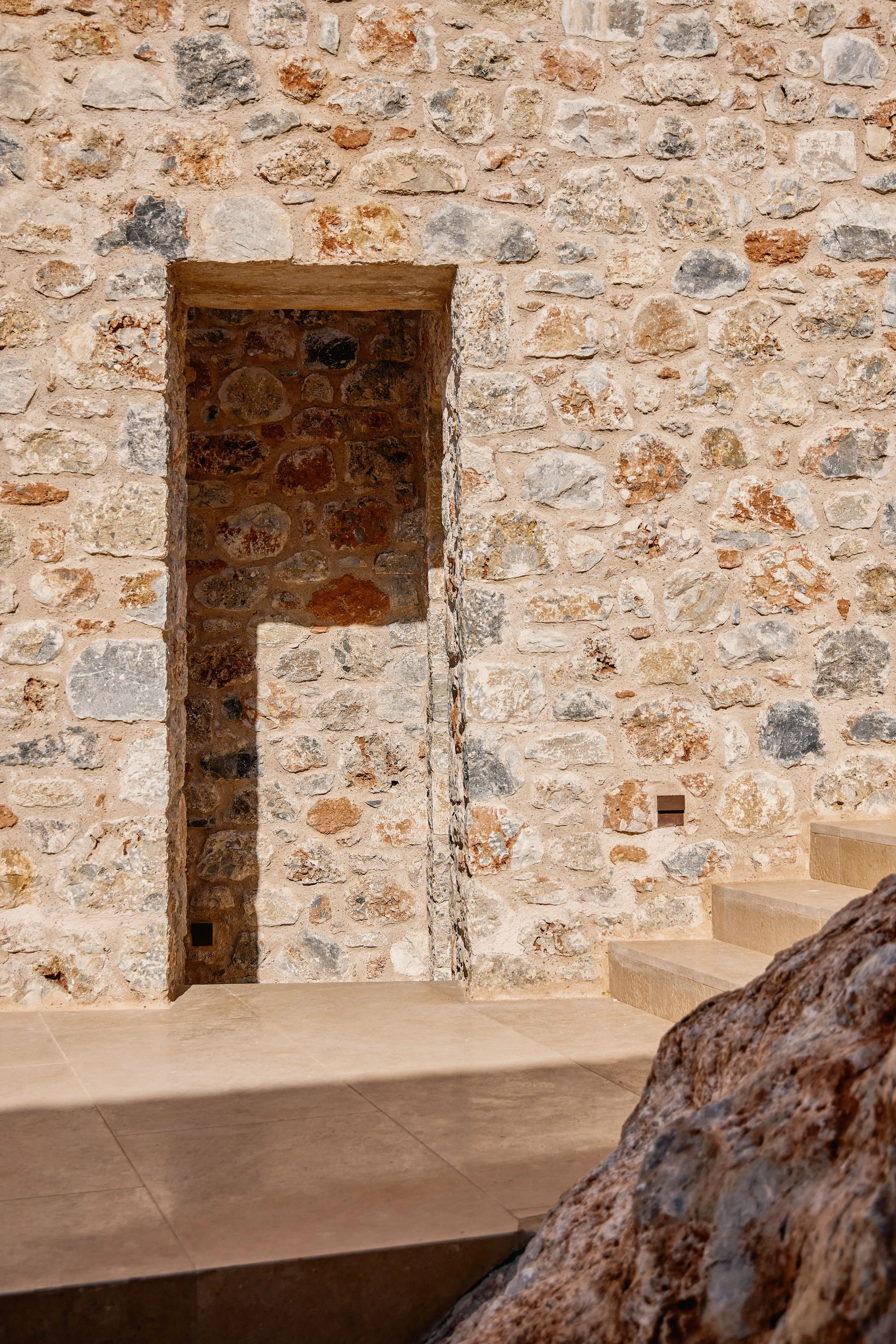 Interior view of a stone wall with steps on the right side and a niche in the wall. The wall is made of irregular stones and the floor is smooth tile.