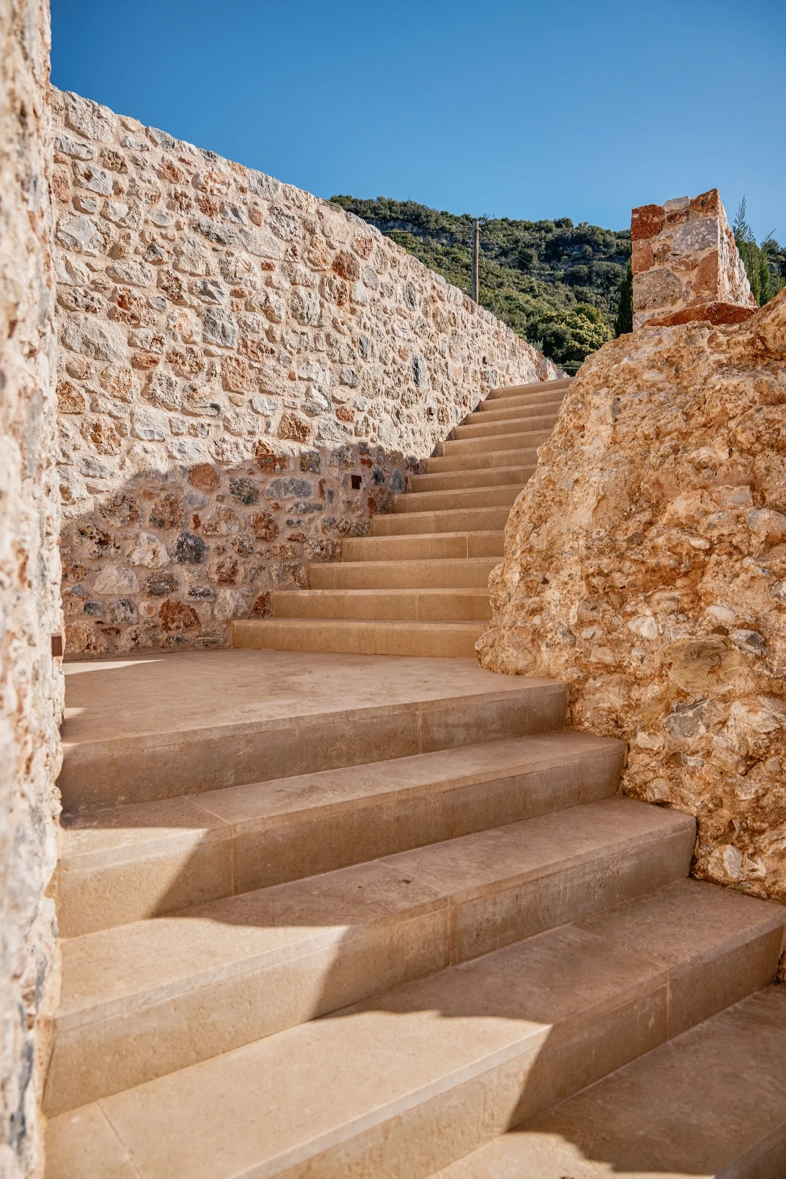 Stone stairs leading upward, flanked by a stone wall and a large rock on the right, with a background of green hillside and a clear blue sky.