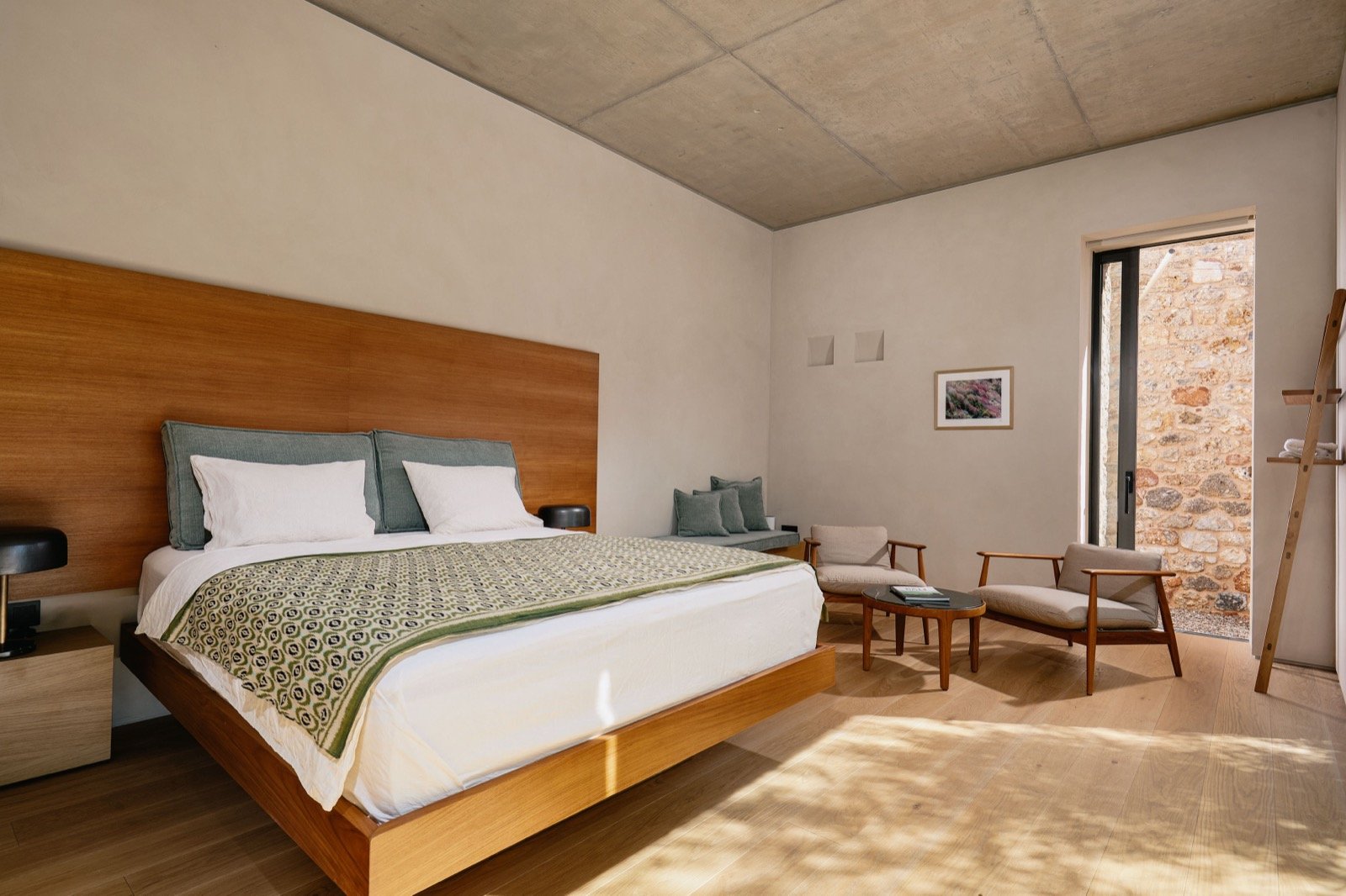 Modern bedroom with a wooden bed frame, green and white bedding, two bedside tables, and a sitting area with two beige armchairs and a small table near a sliding glass door leading outside, with stone wall visible through the door.