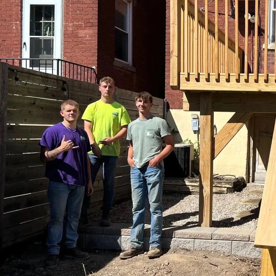 Three young men standing outdoors near a brick building with a wooden deck. They are posing for a photo, with one making a hand gesture. It appears to be a sunny day, and they are in a backyard or construction area.