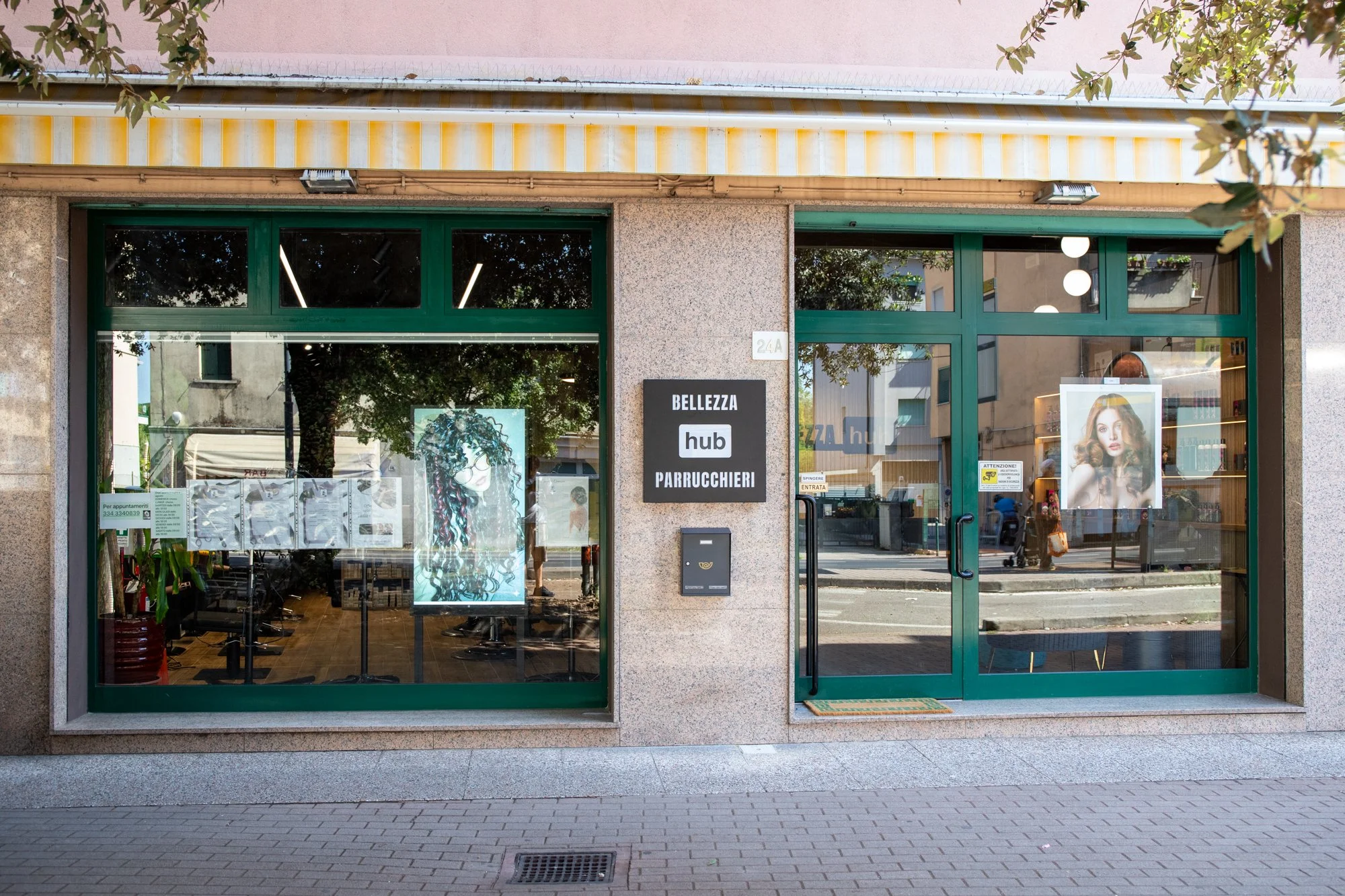 Vetrina di un negozio di parrucchieri con poster di una donna con capelli ricci e un'altra con capelli ondulati, ingresso con porta in vetro, firma 'Bellezza hub Parrucchieri' e dettagli di un'area urbana circostante.