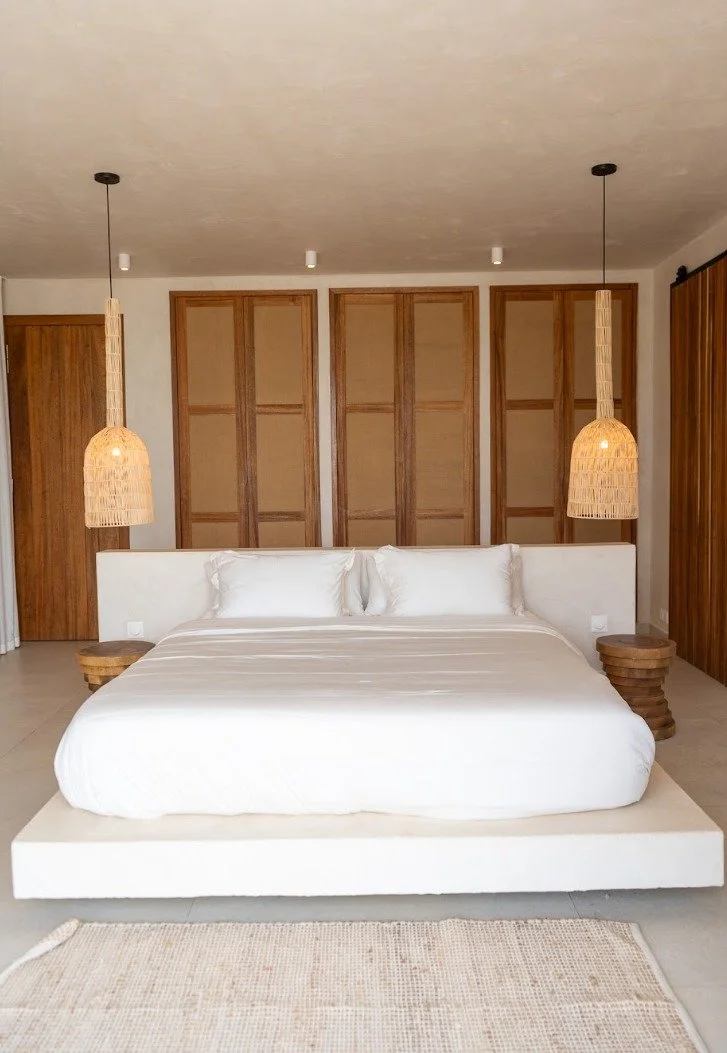 A minimalist bedroom with a king-sized bed, white bedding, two pillows, and a textured beige rug. The room has wooden accents, including two pendant lights on either side of the bed, wooden bedside tables, a headboard, and a wall with wooden framed panels.