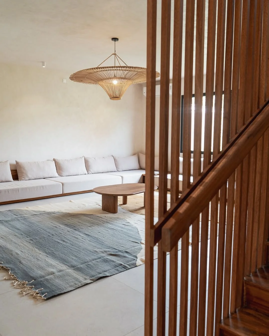 Modern living room with a long cream-colored sofa, a round wooden coffee table, a woven pendant light, a large area rug, and a wooden staircase with vertical slats.