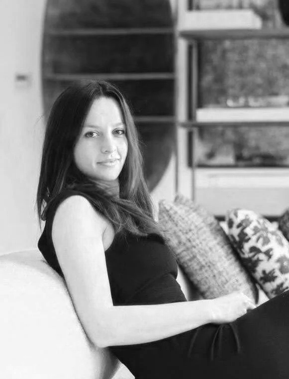 A woman with long dark hair sitting on a sofa in a room with shelves and pillows in the background.