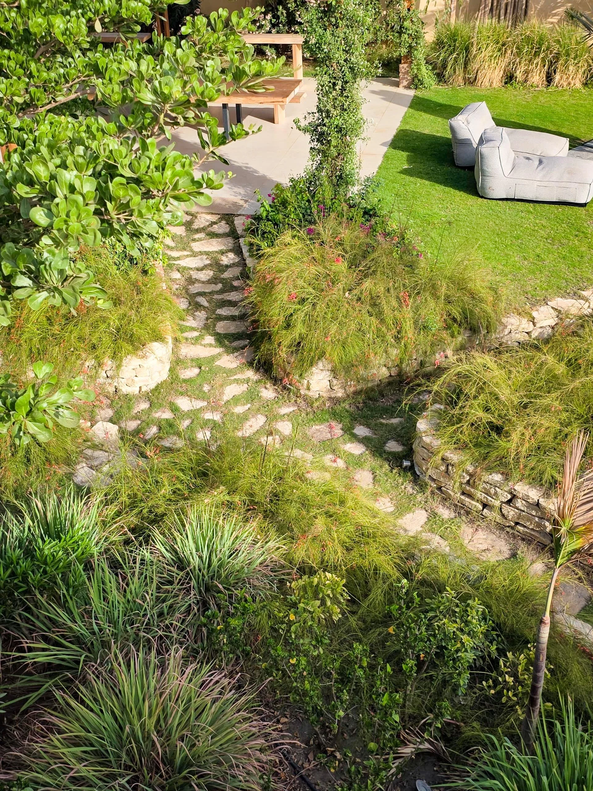 A lush garden with various green plants, a stone pathway, a patio with benches, and outdoor furniture including two armchairs facing away.
