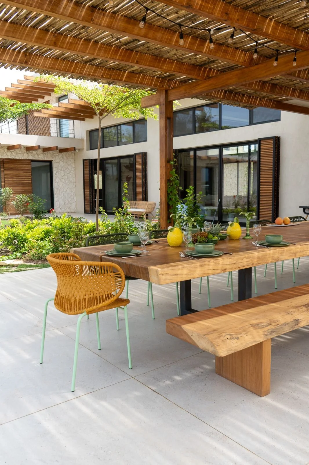 Outdoor patio dining area with a wooden table set with green plates, glasses, and yellow squash-shaped vases, surrounded by chairs, with a modern house and garden in the background.