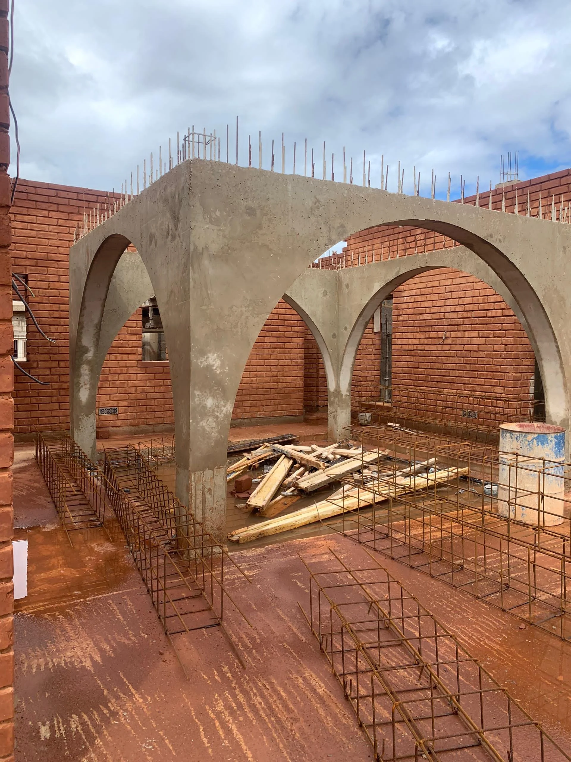 Building construction site with concrete arches and brick walls under a cloudy sky, with metal rebar and construction materials on the ground.