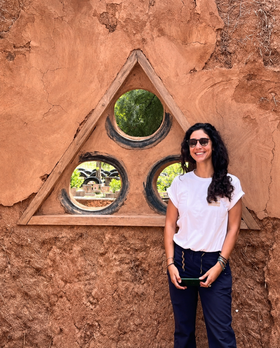 A woman with curly dark hair wearing sunglasses, a white t-shirt, and dark pants, smiling while standing outdoors next to a triangular wooden frame with three circular cutouts in a dirt wall, with trees and tires visible in the background.