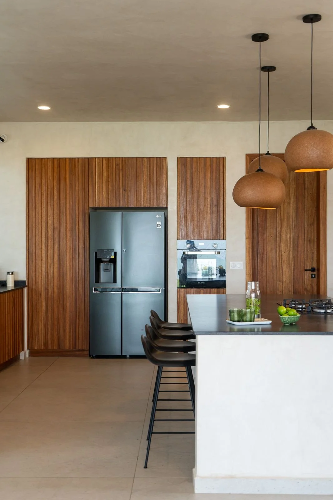 Modern kitchen with wooden cabinets, stainless steel LG refrigerator, wall oven, black bar stools, white island with dark countertop, hanging pendant lights, and green apples on the counter.