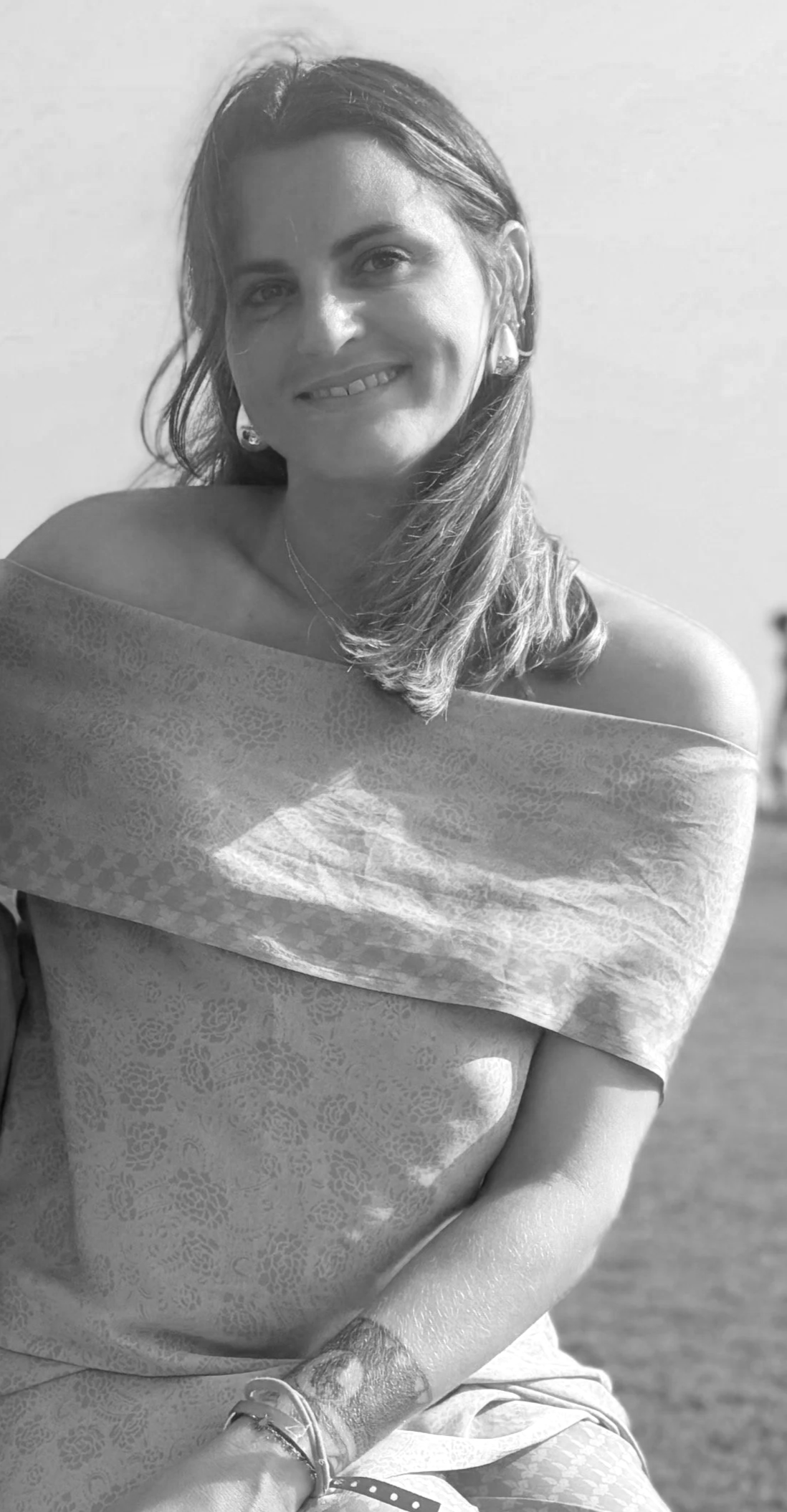Black and white photo of a smiling woman sitting outdoors wearing a patterned off-the-shoulder dress, with medium-length hair, earrings, and bracelets on her wrist.