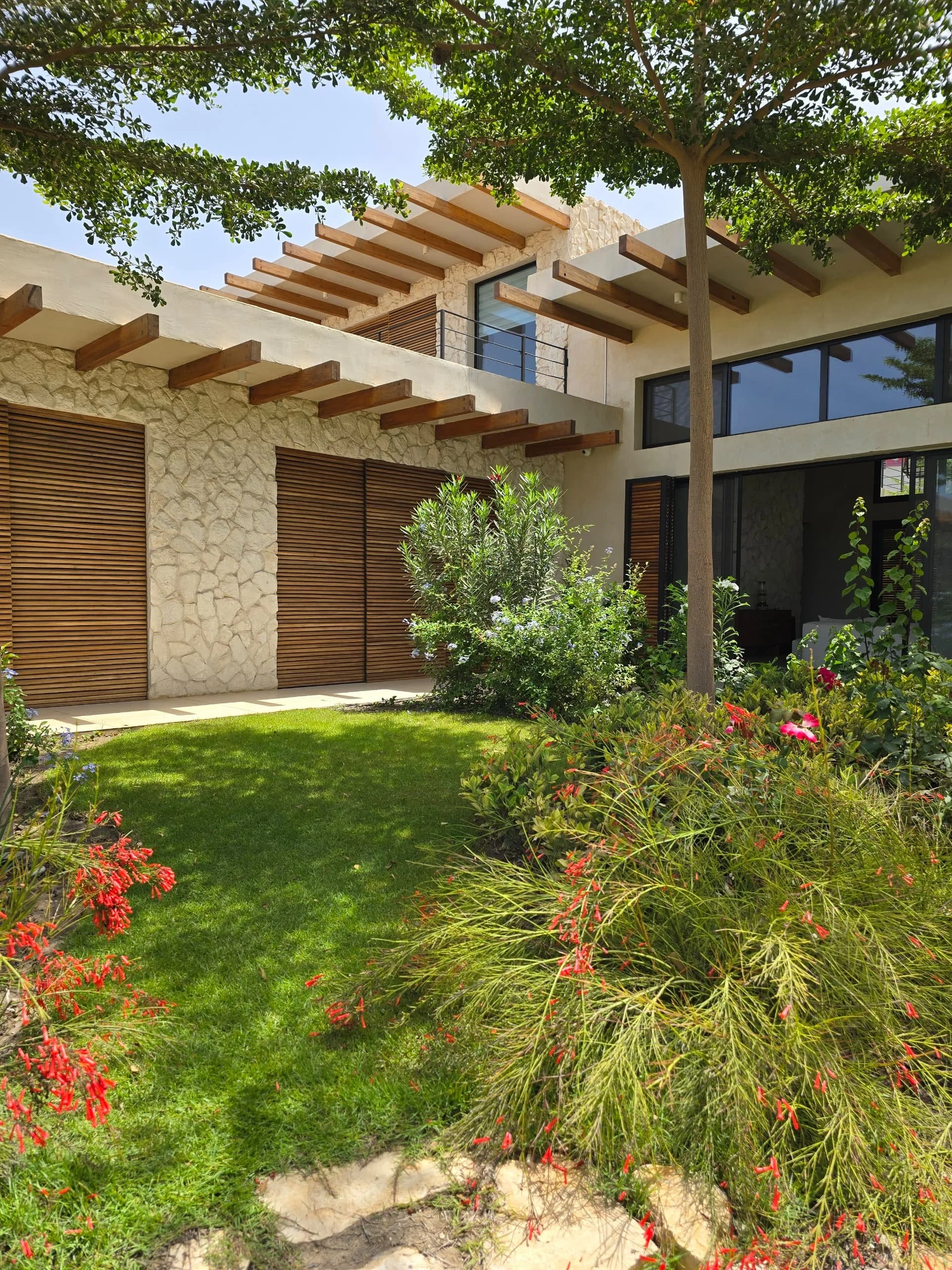 A modern house with stone and wood exterior, large windows, and surrounded by lush greenery and colorful flowering plants in a sunny setting.