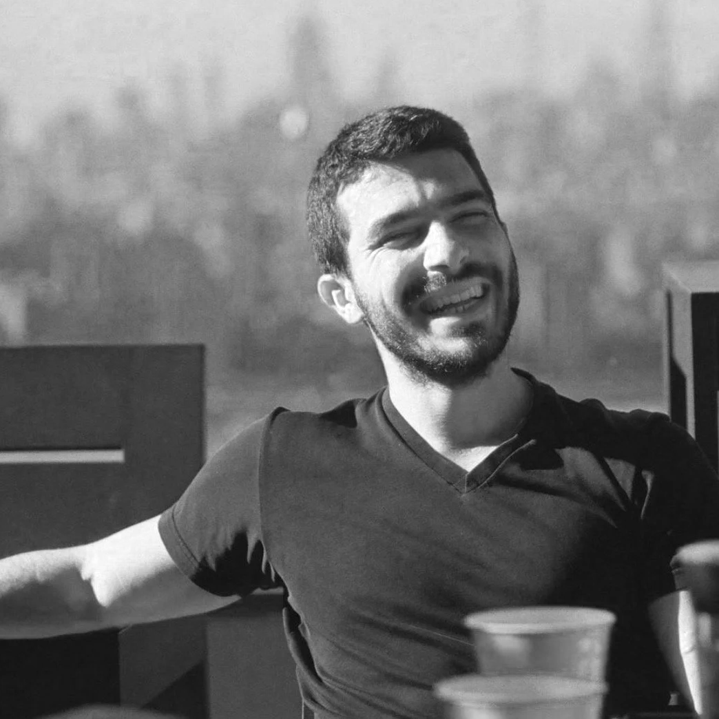A smiling man with a beard and short dark hair, wearing a dark t-shirt, sitting at a table outdoors with sunlight, possibly during daytime.