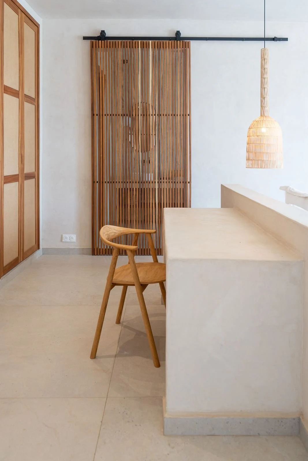 A minimalist living room featuring a wooden chair, a beige island or counter, a rattan hanging pendant light, a wooden sliding room divider, and a wall with an electrical outlet.