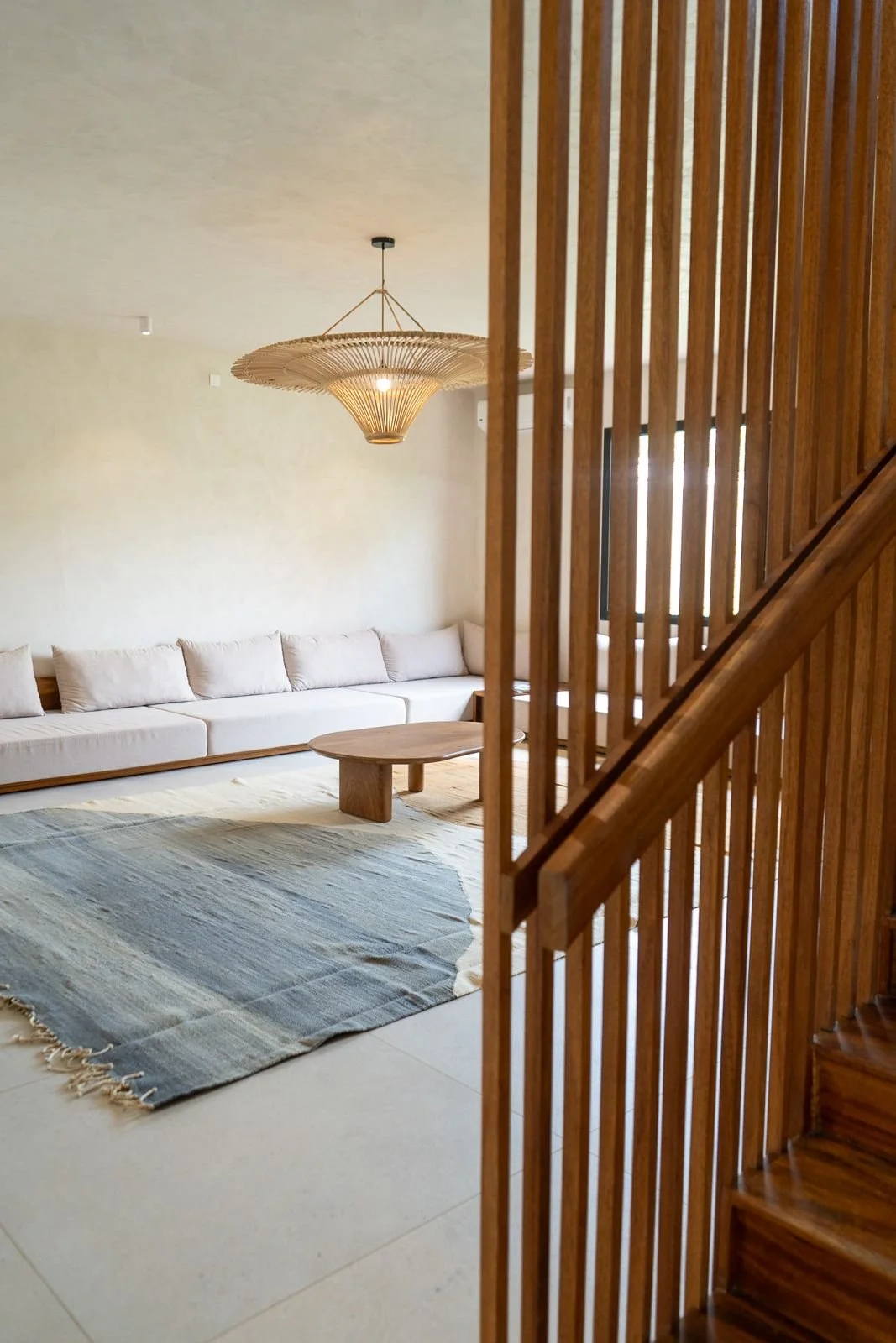 Modern living room with a white sofa, a round wooden coffee table, a woven light fixture, a textured rug, and a wooden staircase railing.
