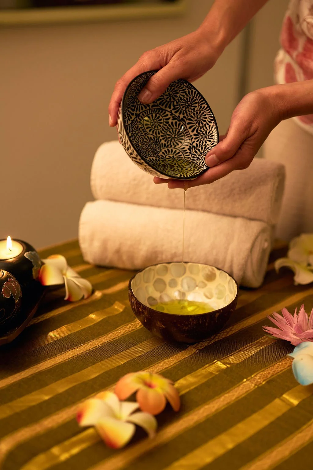 Une personne verse de l'huile dans un bol décoratif. Il y a des bougies et des fleurs autour, suggérant une ambiance zen ou de massage. Le tapis a des rayures dorées.