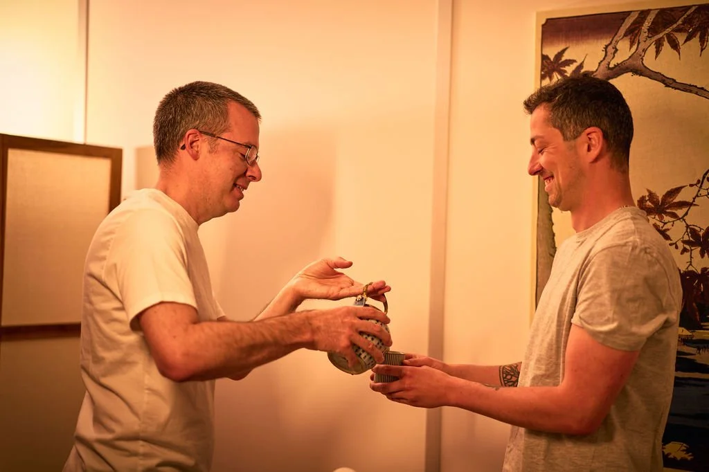 Deux hommes souriants échangent de l'argent dans une pièce. L'homme de gauche porte des lunettes et une chemise claire, l'autre un t-shirt gris clair. Une décoration murale est visible en arrière-plan.