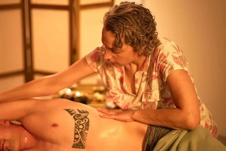 Une femme donne un massage à une personne allongée, visible partiellement, dans une ambiance chaude et intime.