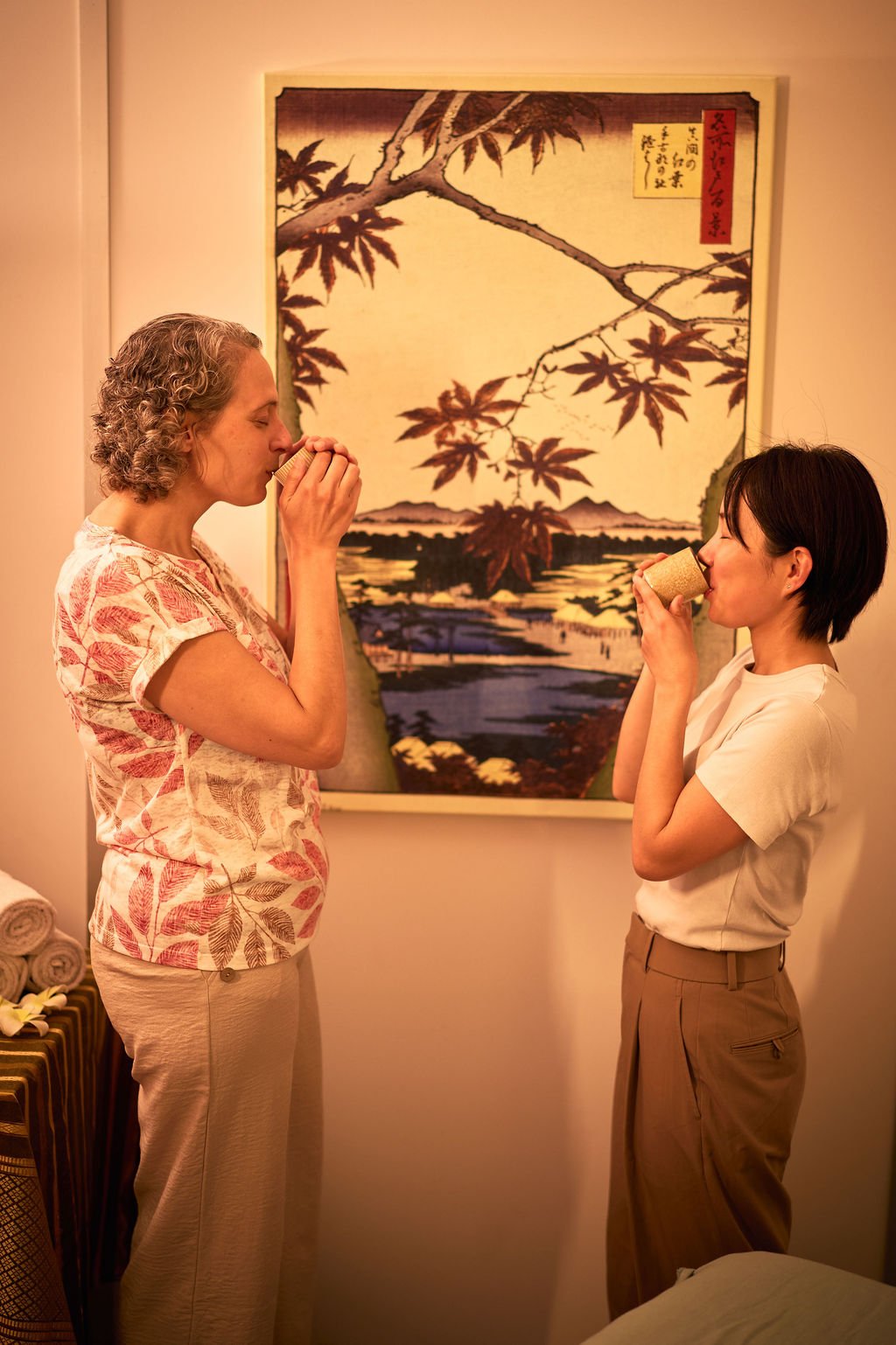 Deux femmes boivent une boisson dans une pièce avec une grande œuvre d'art japonaise en fond. Une femme porte un haut à motifs feuillages et l'autre une chemise blanche, et elles tiennent des tasses en mains.