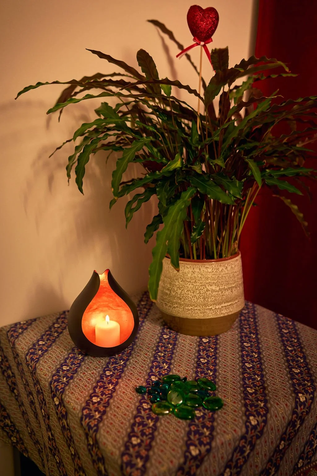 Une plante verte dans un pot beige à motifs, un porte-encens avec une bougie allumée à côté, et des pierres vertes sur une table recouverte d'une nappe à motifs.