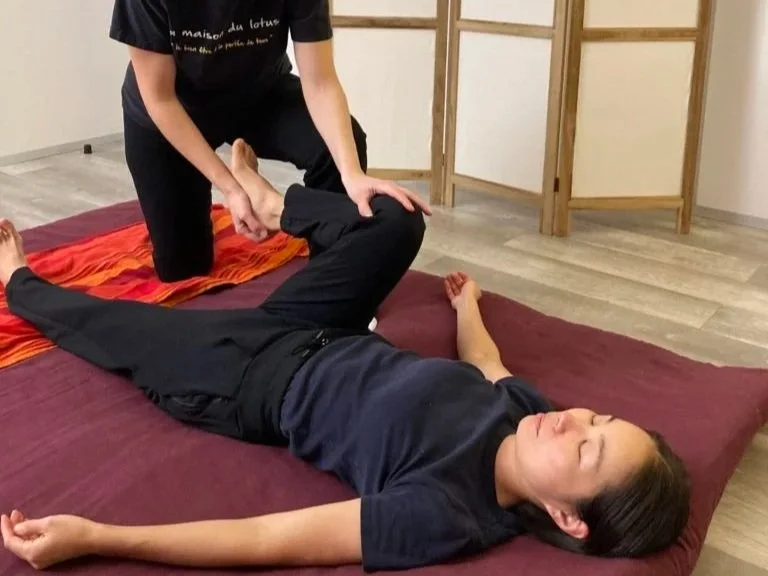 Une personne reçoit une thérapie de relaxation ou de physiothérapie sur un tapis avec un thérapeute. La personne est allongée sur le dos, les yeux fermés, dans une pièce calme et sobre.
